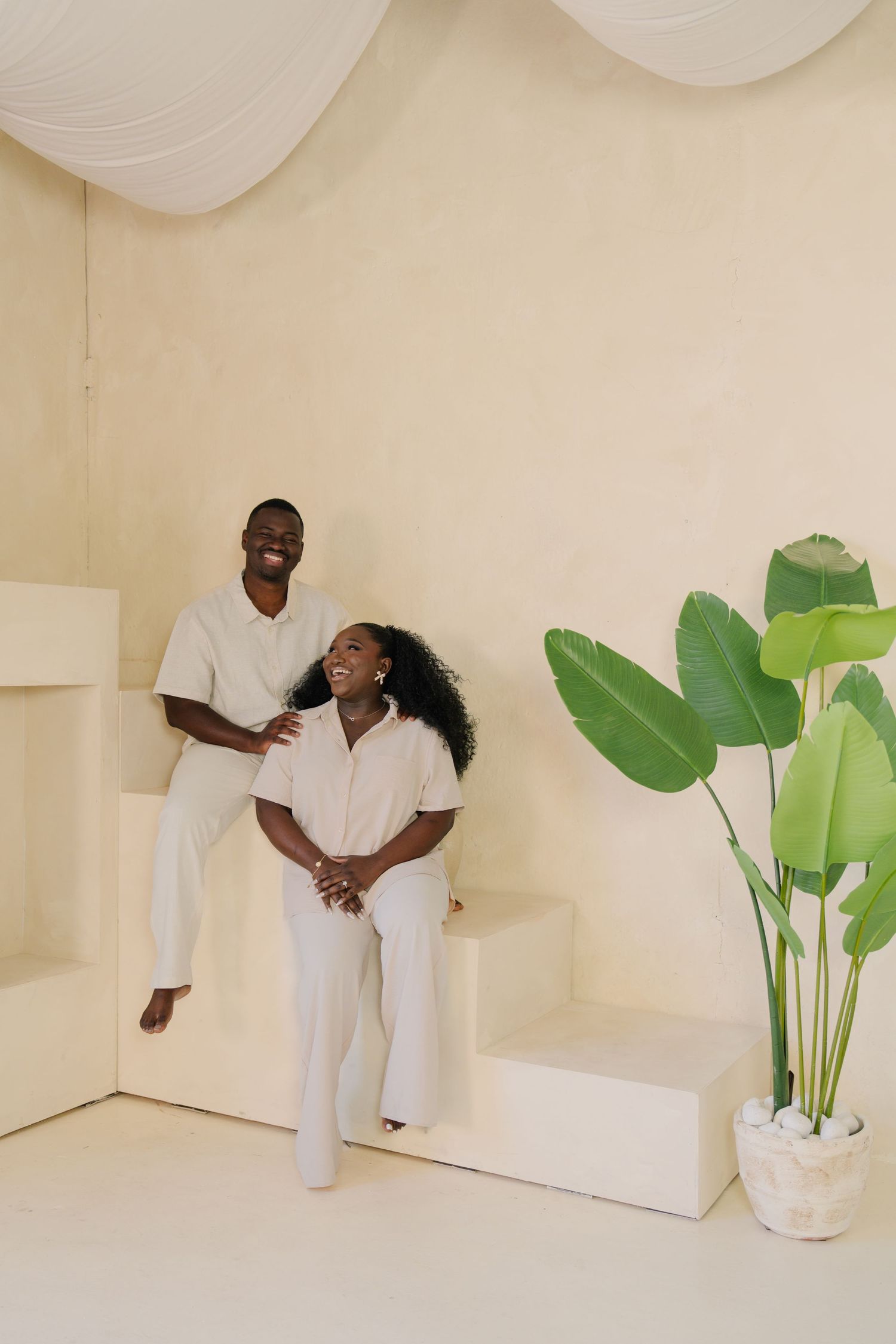 Happy Black couple in white outfits laughing together on white steps beside a tropical plant in a bright minimalist room.