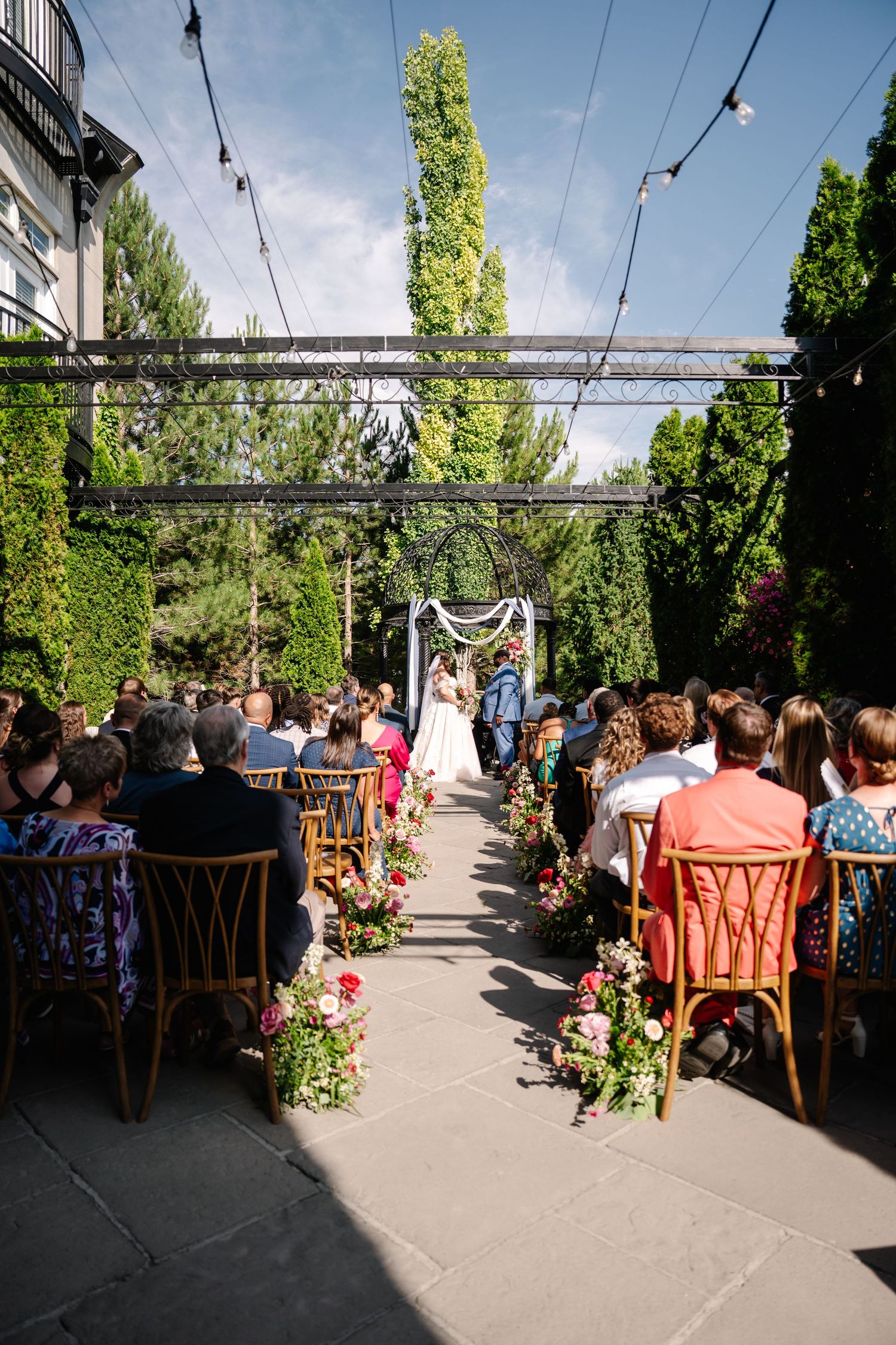 Outdoor wedding ceremony setup with string lights overhead and rows of wooden chairs along a flower-lined aisle.