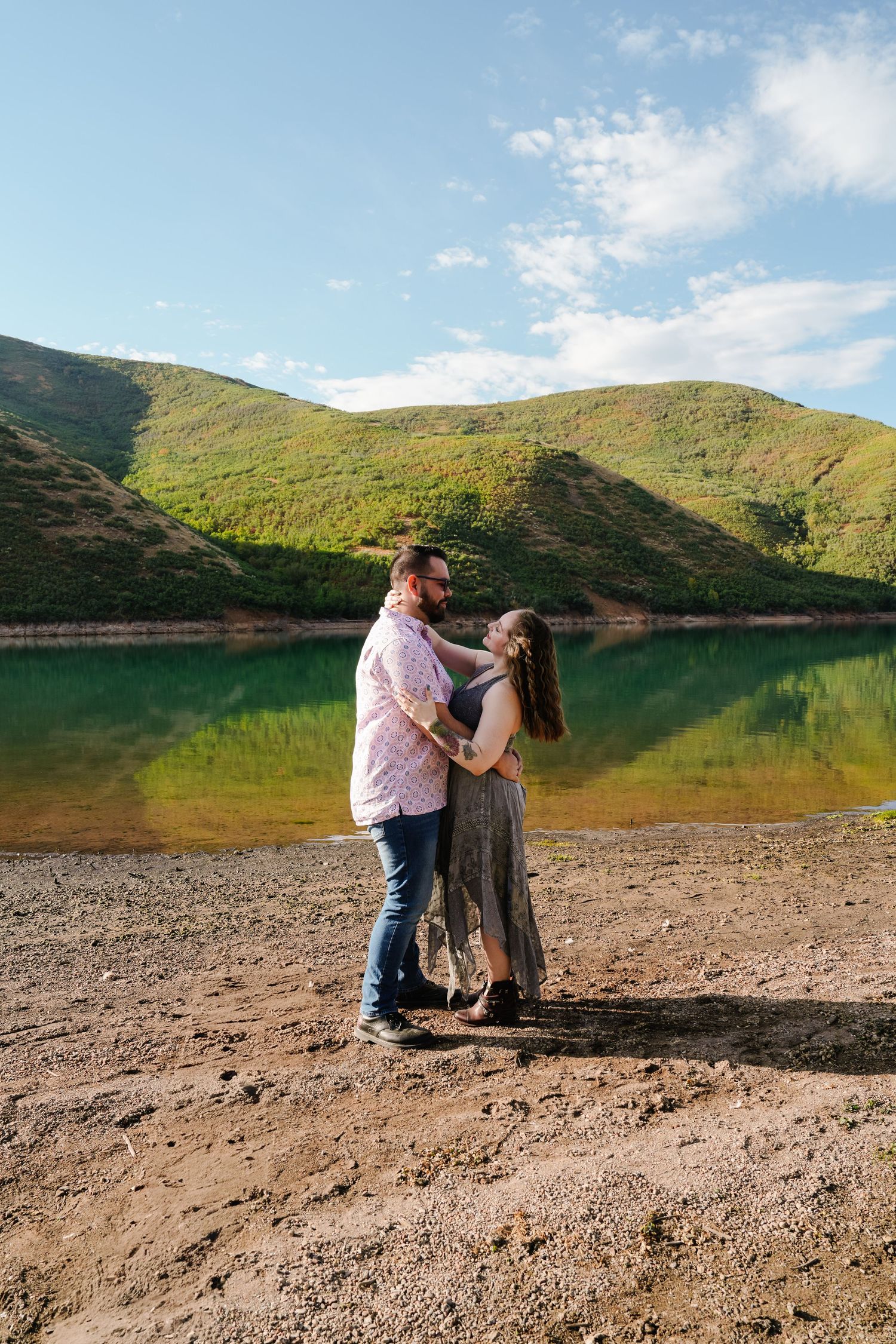 Couple dancing on a shoreline with rolling hills and reflective lake waters behind them.