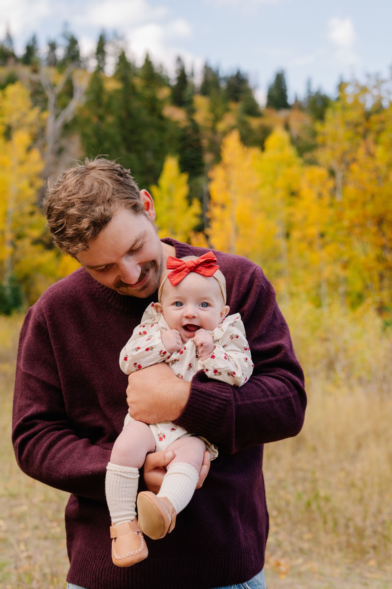 Father in burgundy sweater holds smiling baby girl with red bow amid golden autumn aspen trees.