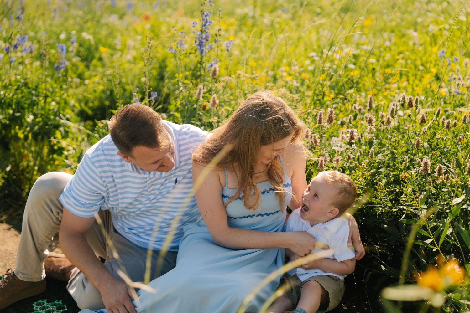 Family of three sitting in a sunny wildflower meadow, parents smiling at laughing baby on a warm summer day.