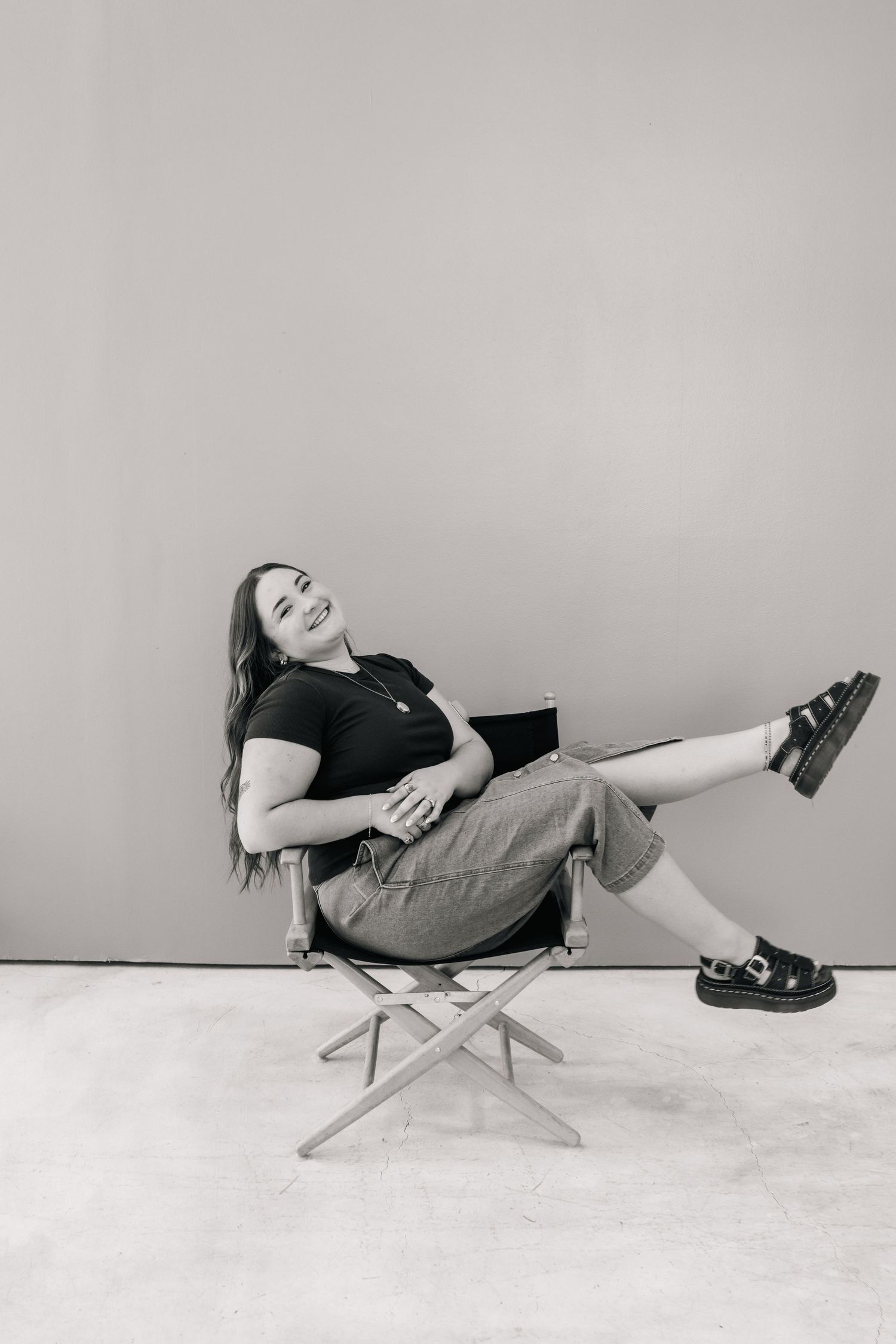 A smiling young woman relaxes in a folding director's chair, wearing a black top and jeans, in a black and white photo.