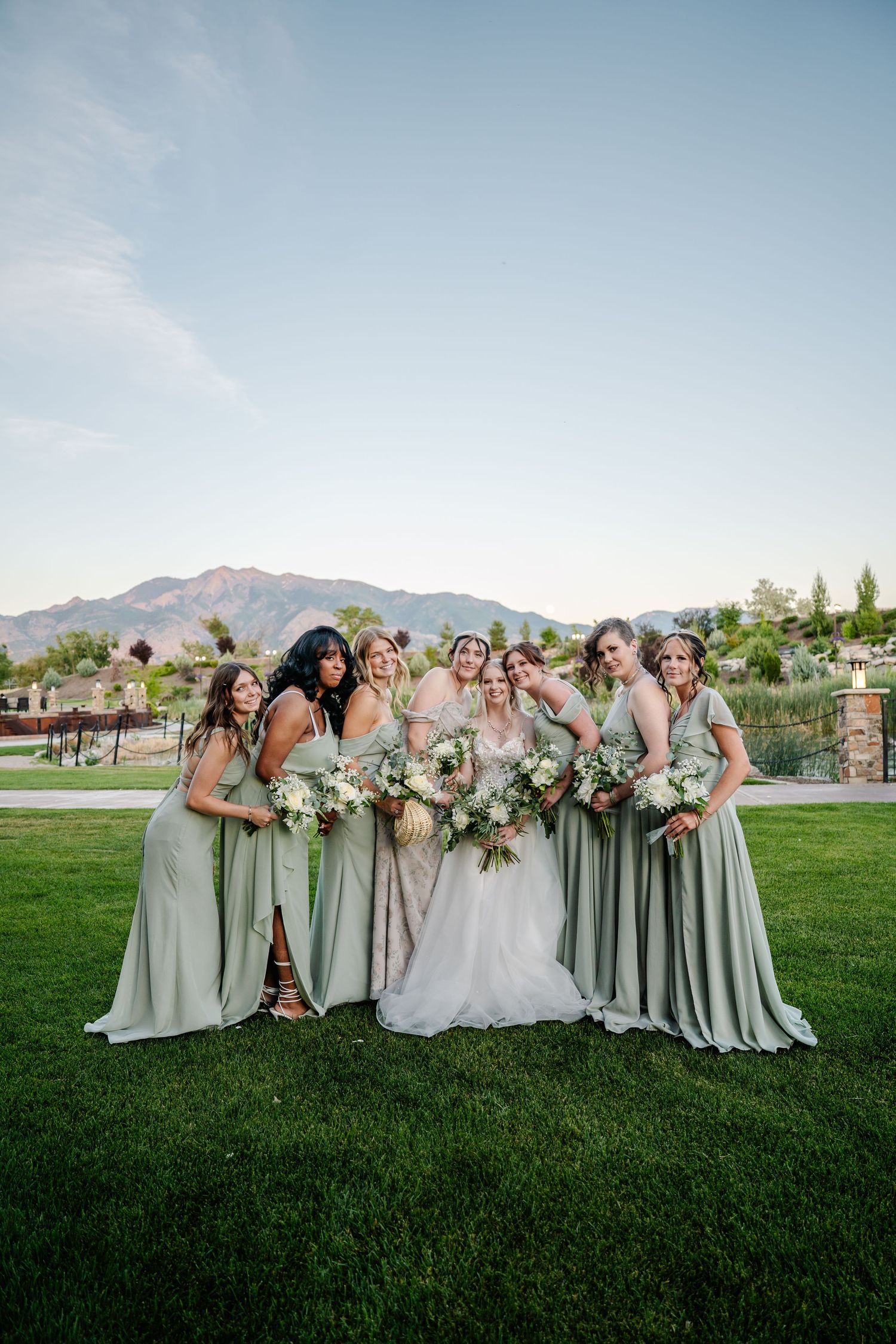 Bride in white gown poses with six bridesmaids in sage green dresses holding bouquets on a lawn with mountain views.