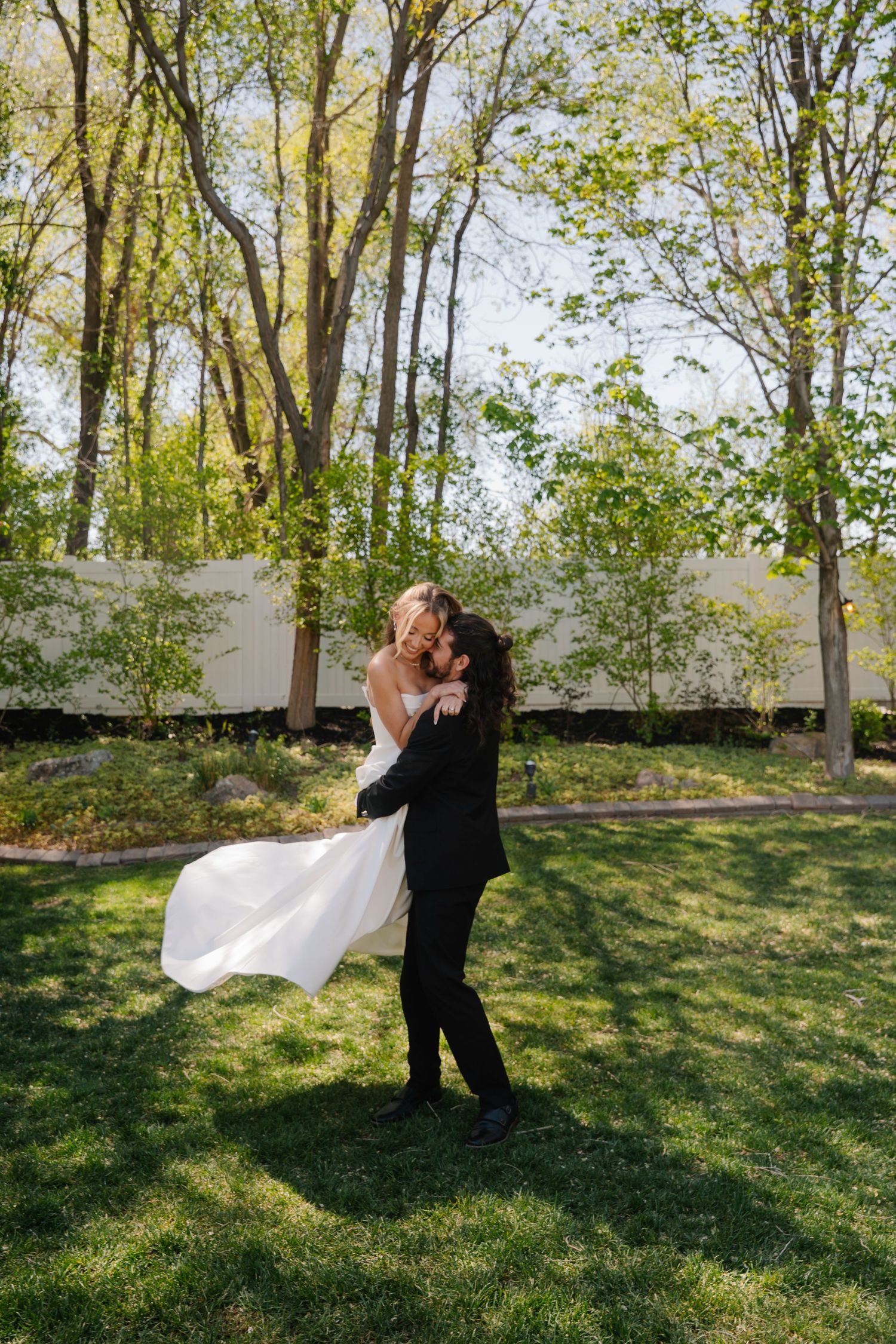 Groom lifting bride in white dress outdoors on green lawn surrounded by trees on their wedding day.
