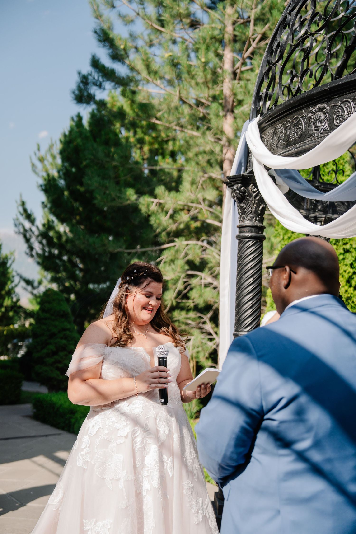 Wedding couple exchanging vows under an ornate metal gazebo decorated with white fabric.