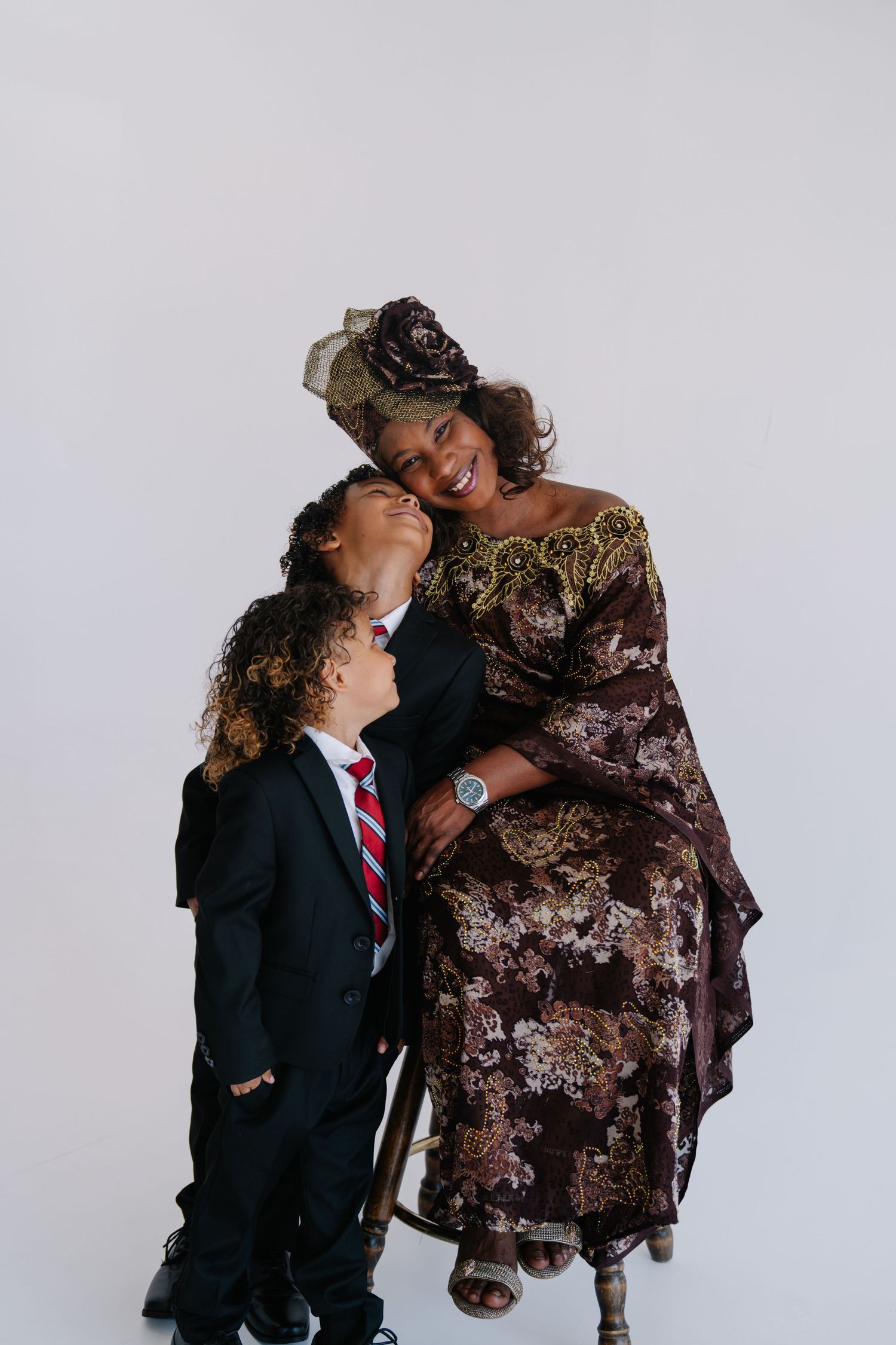 A smiling woman in ornate African attire embraces a young boy in a black suit and red tie against a white background.