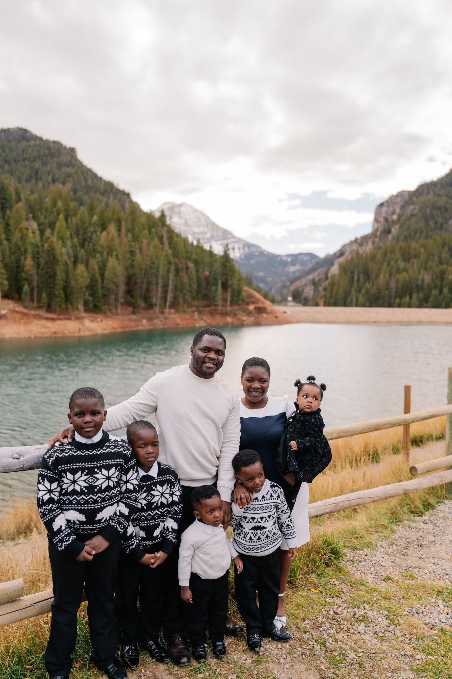 Family of six posing by a mountain lake surrounded by pine trees on a cloudy day.