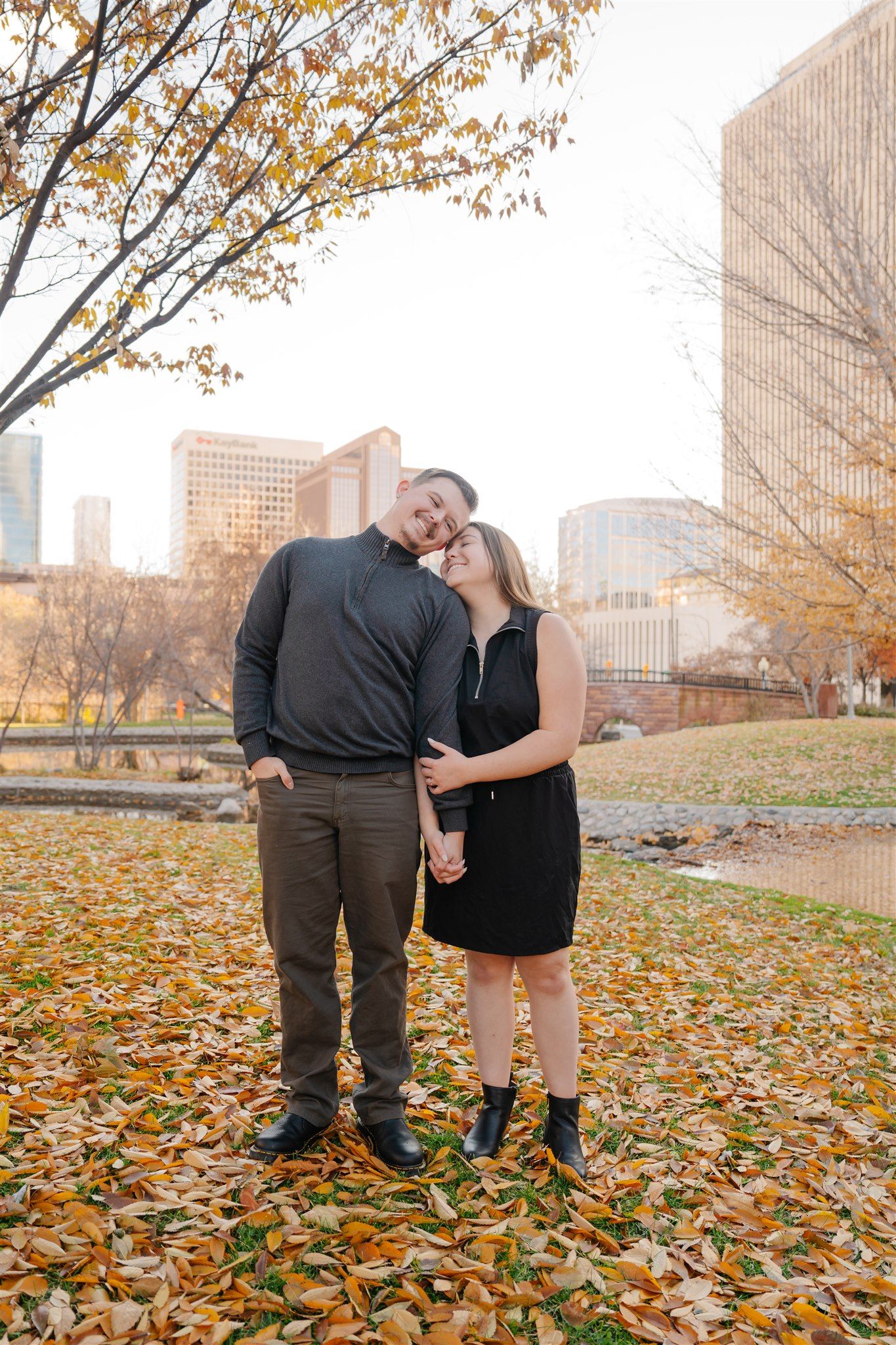 Couple posing together in a park during autumn with fallen leaves and city buildings in background.