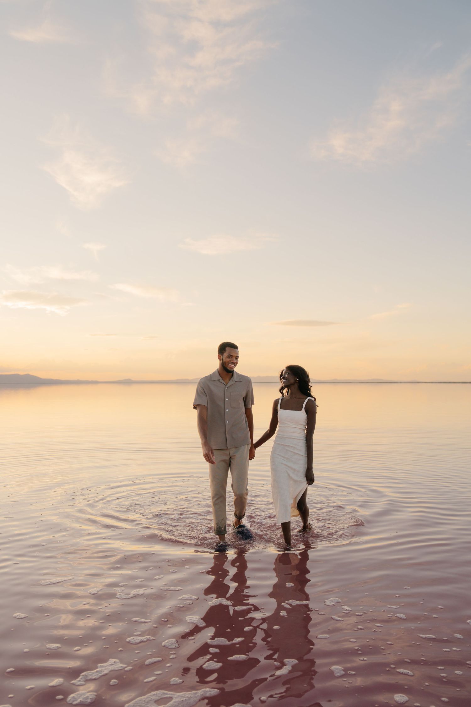 A couple stands together in shallow pink-tinted water during sunset at a salt lake.