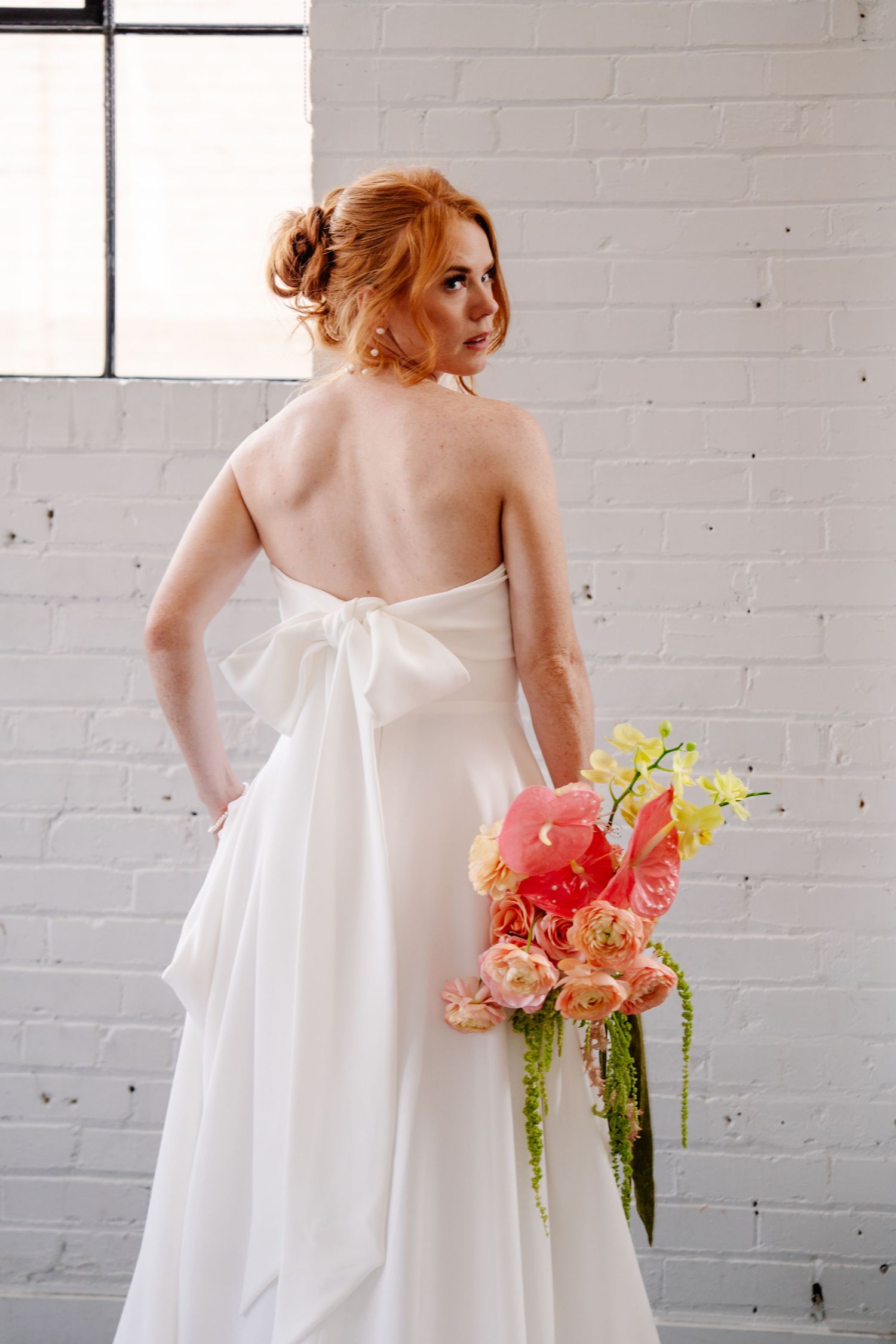 A flowing white wedding dress with a bouquet of colorful flowers against a white brick wall.