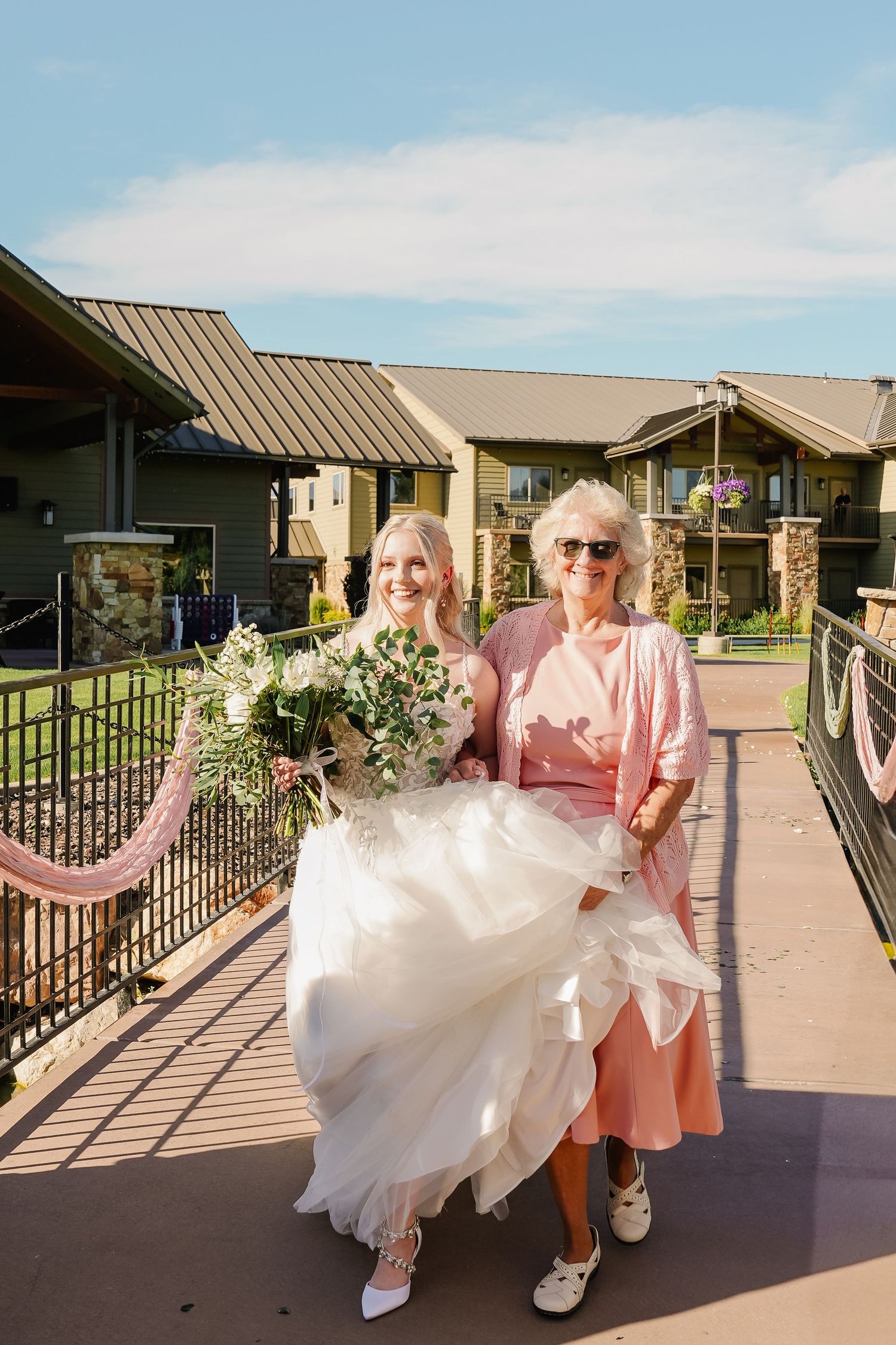 Wedding dress being carried along outdoor walkway with pink-clad helpers.