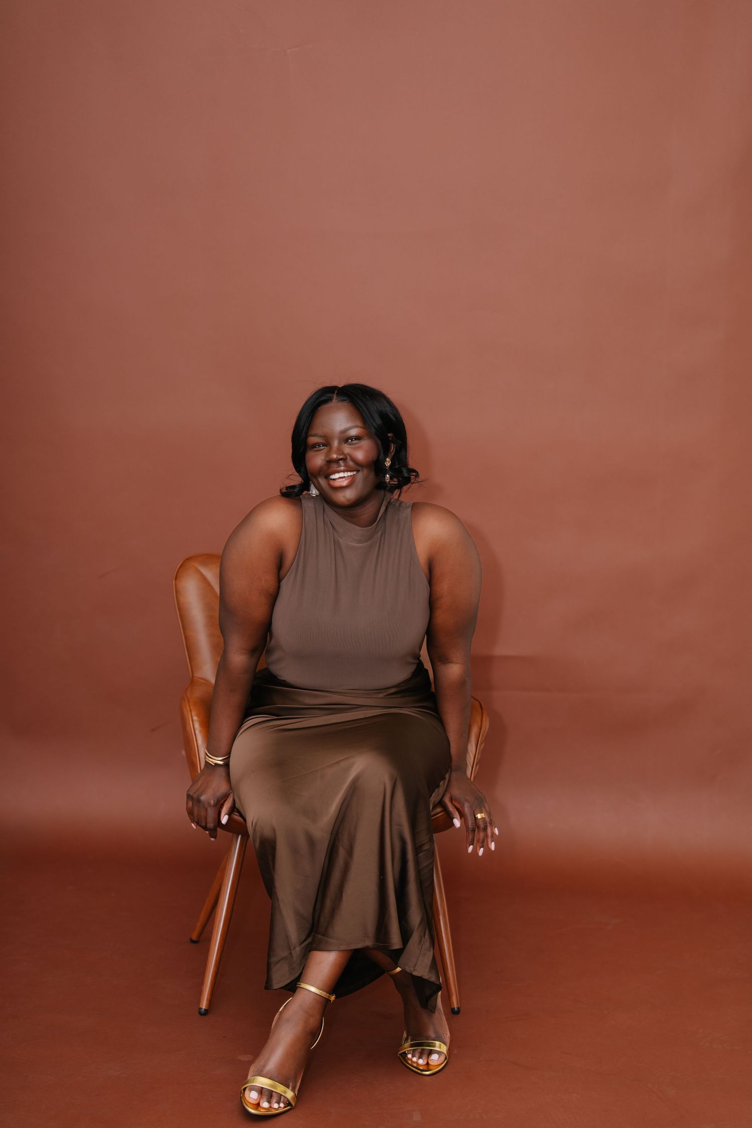 A smiling woman in an olive green dress and gold heels sits confidently in a wooden chair against a warm brown backdrop.