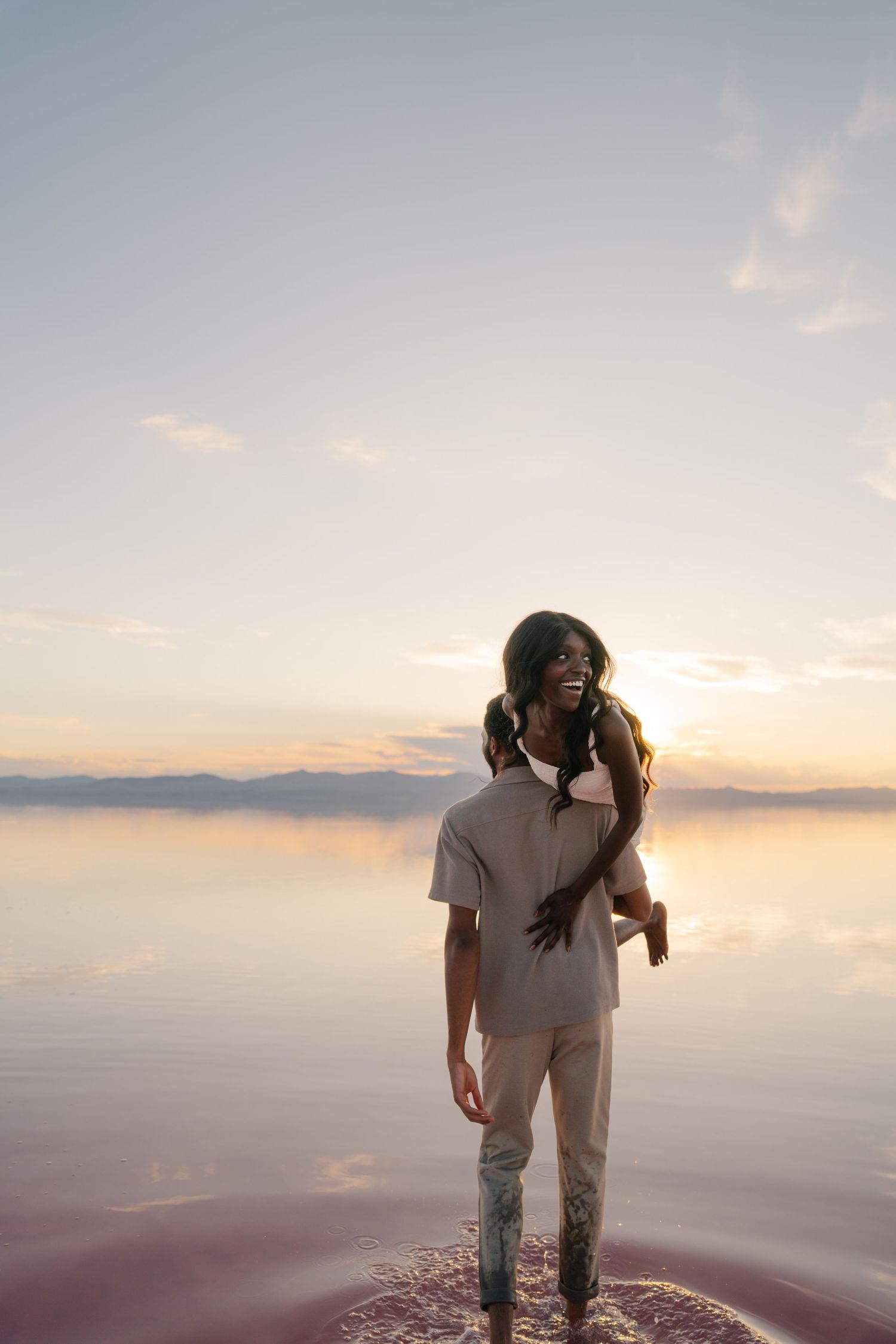 A couple in white outfits embraces while standing in shallow water during sunset.