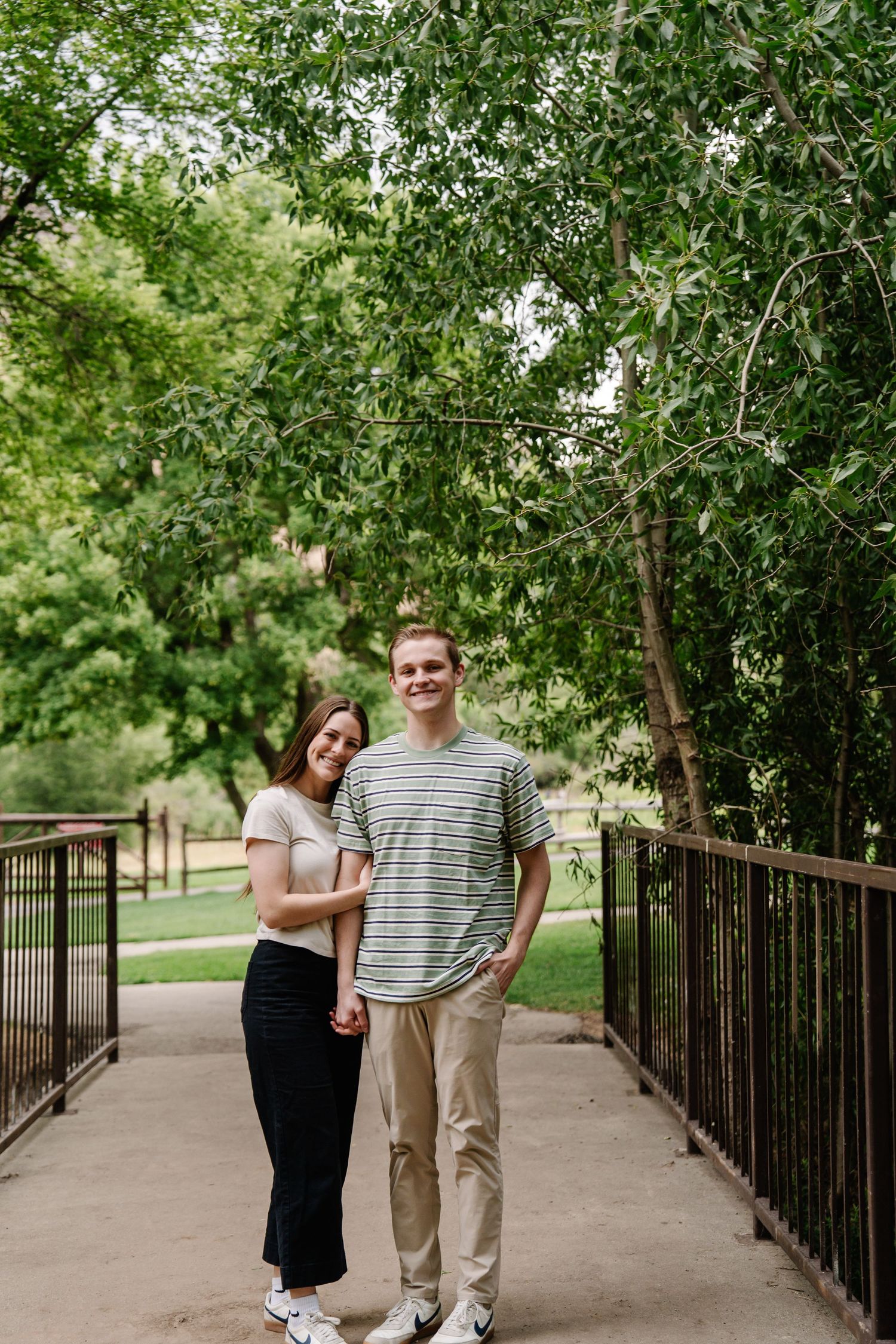 A couple standing on a bridge pathway surrounded by lush green trees.