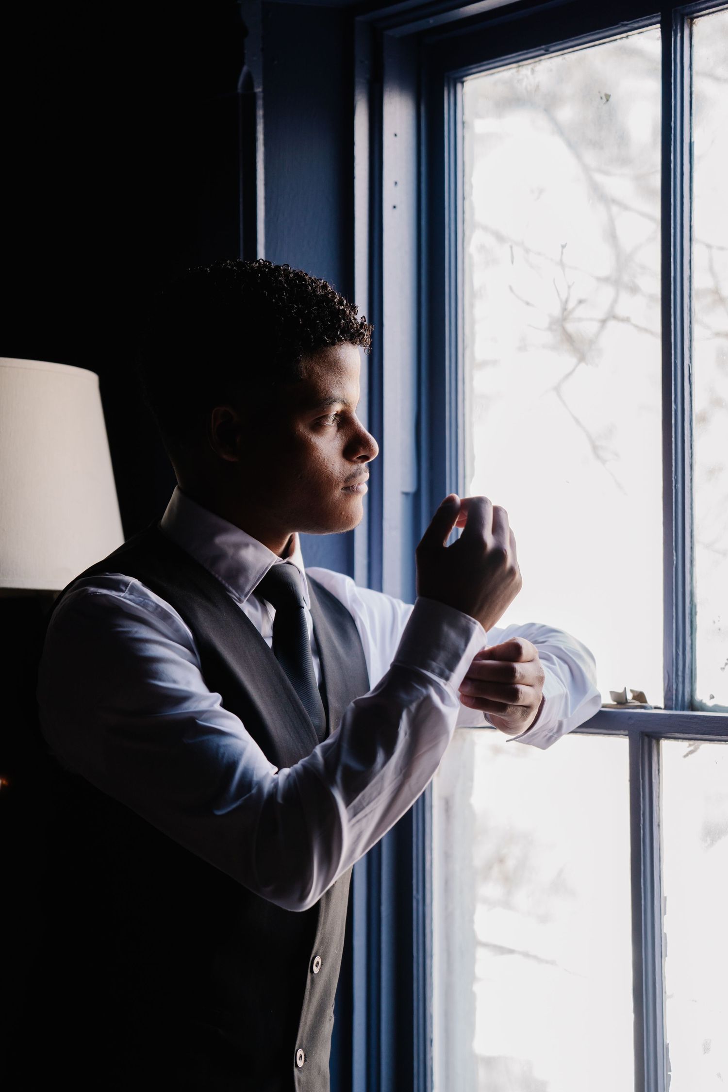 A well-dressed groom adjusts his cufflinks by a bright window, wearing a gray vest and tie on his wedding day.
