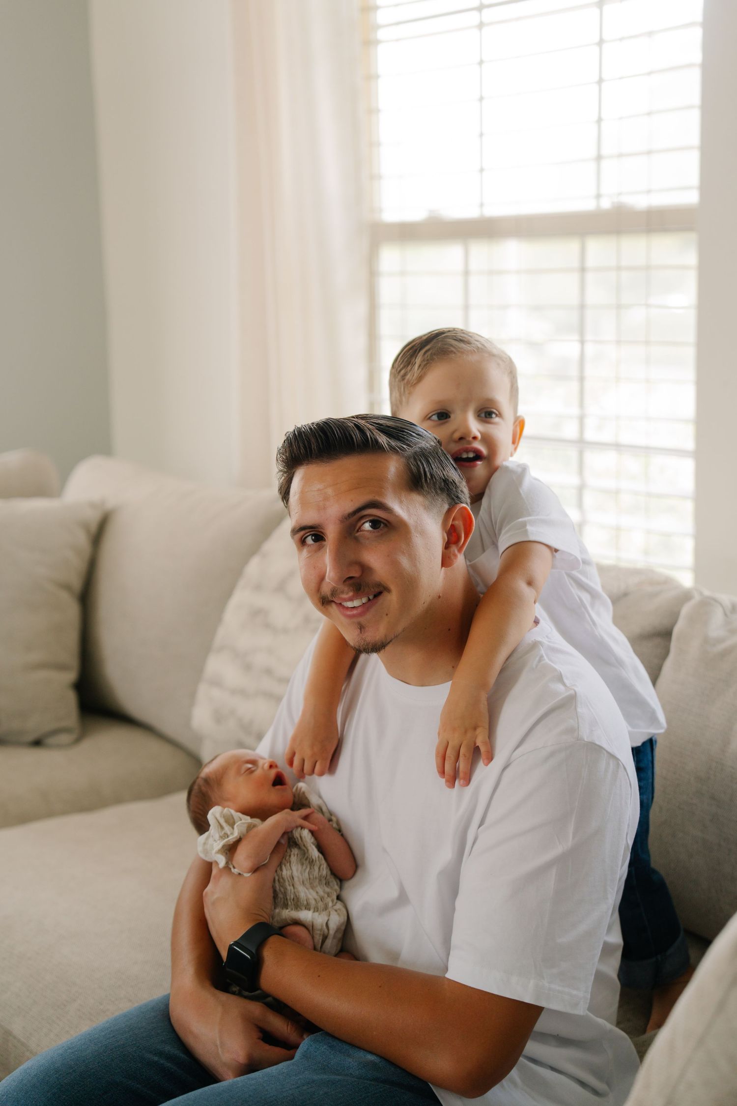 A smiling father holds a newborn while his toddler son sits on his shoulders on a couch.