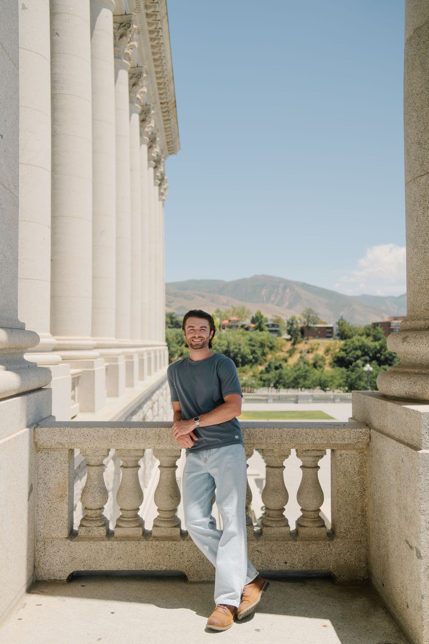 Man in gray t-shirt and jeans leaning on stone balustrade of neoclassical building with mountain views.