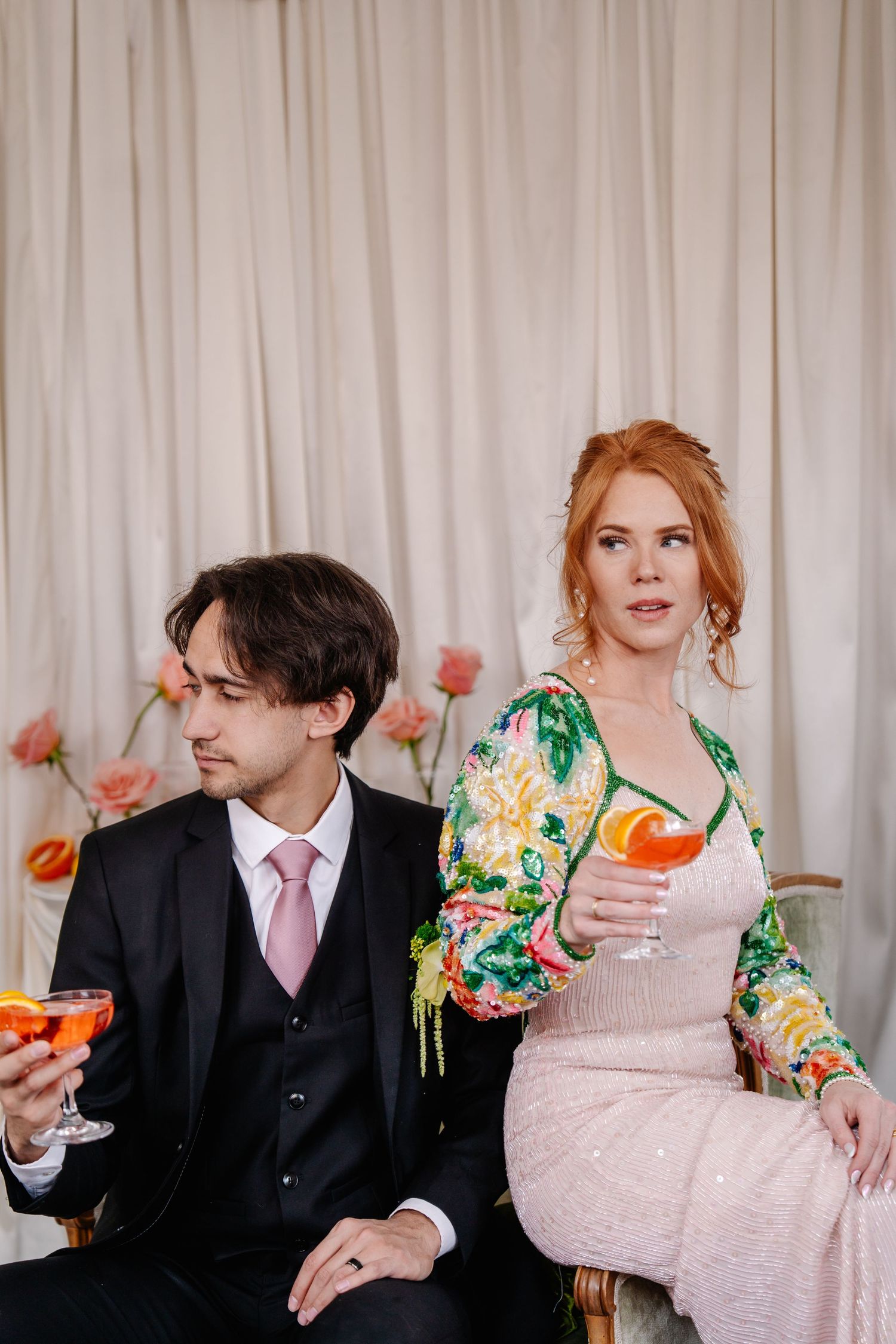 Wedding couple in formal wear enjoying drinks and laughter against white curtain backdrop.