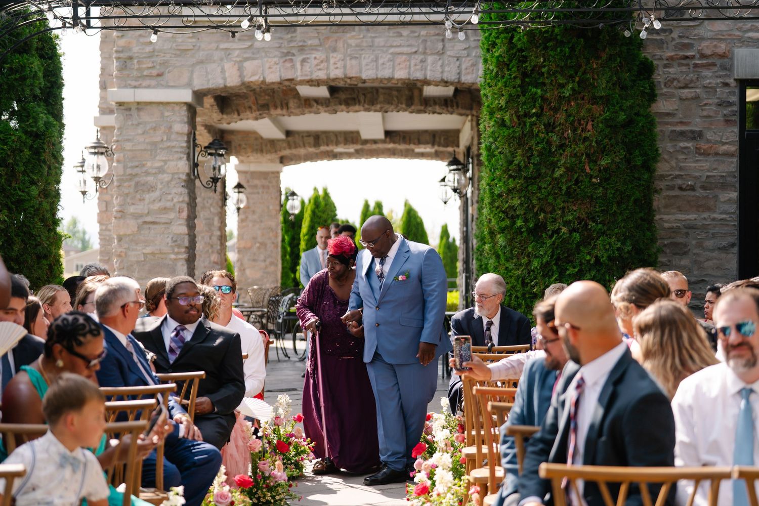 Wedding ceremony guests seated in rows under a stone archway decorated with flowers.