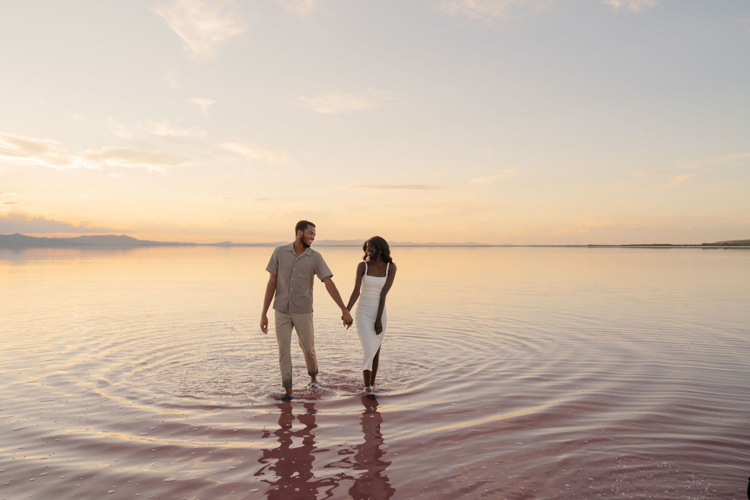 Silhouetted couple walking in shallow water during sunset creating ripples on the calm surface.