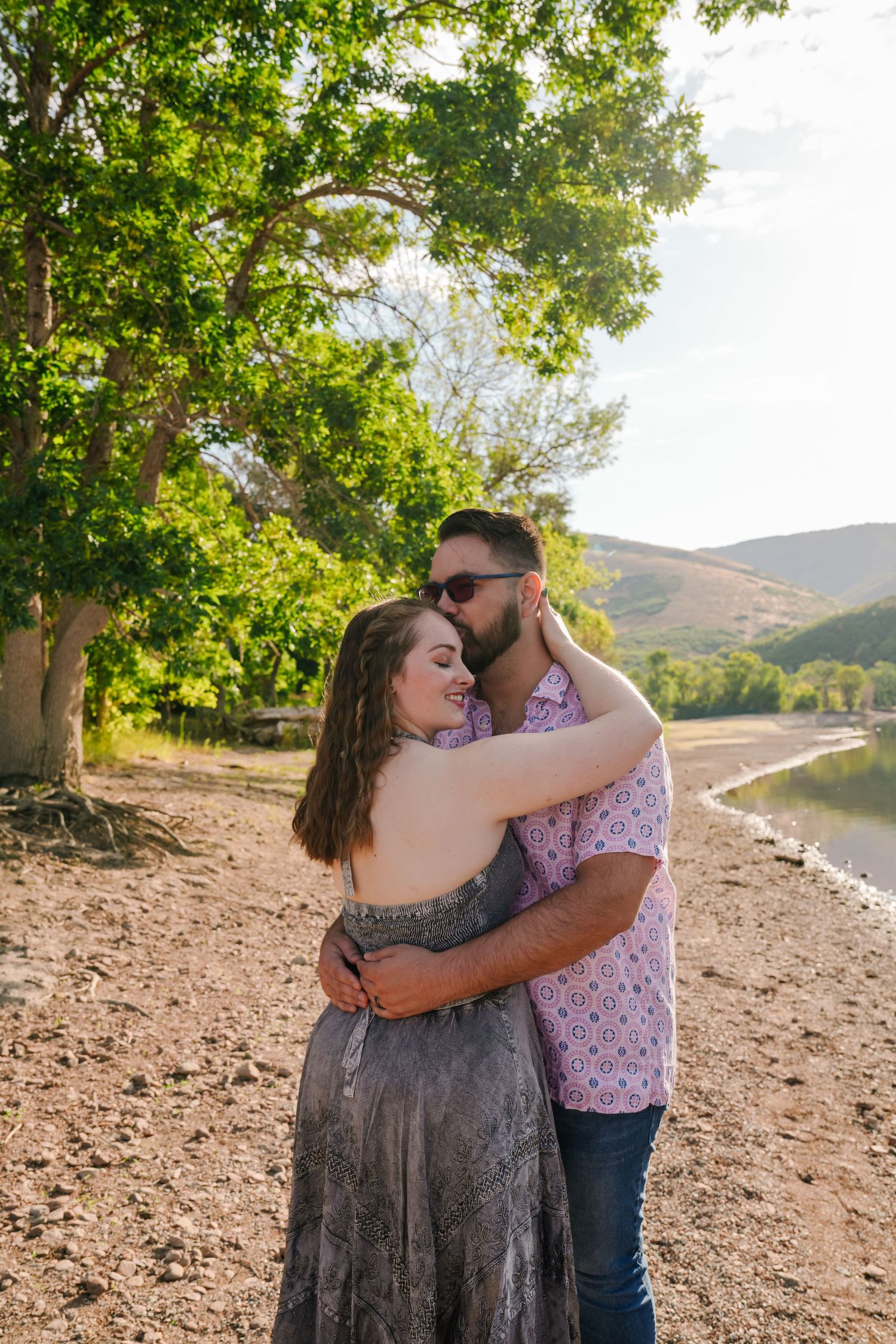 A romantic couple embraces on a lakeside shore with green trees and mountains in the background.