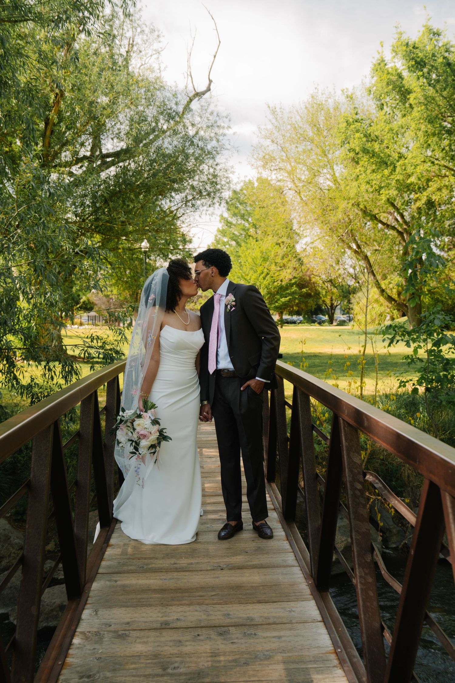 Bride and groom kissing on a wooden bridge surrounded by lush green trees on their wedding day.