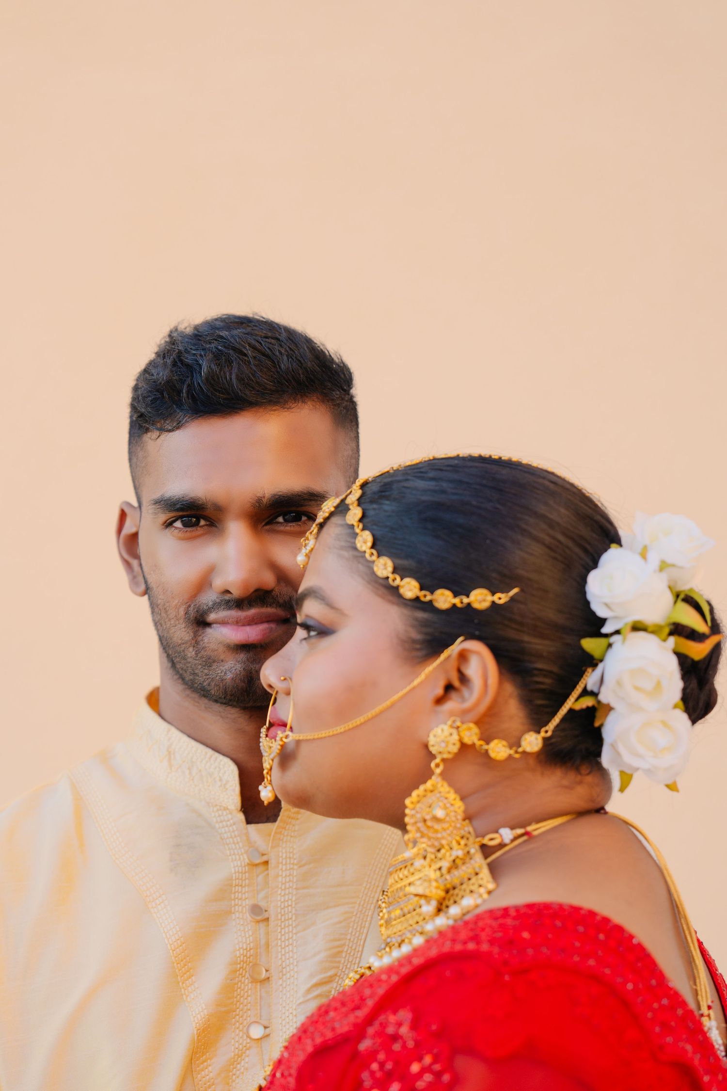 A couple in traditional South Asian wedding attire with gold jewelry and white flowers against a peach background.