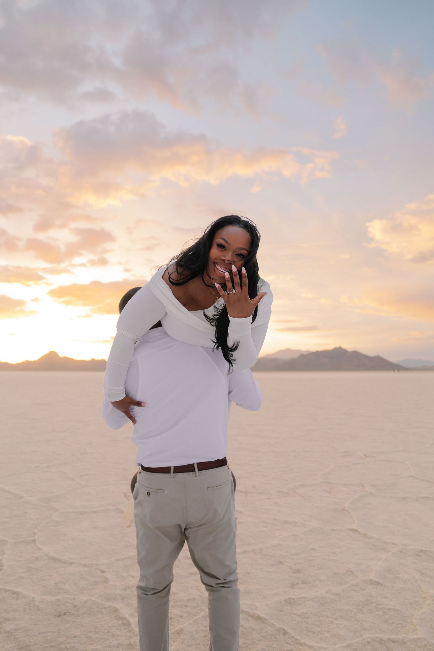 A joyful woman shows off her engagement ring while being carried by her partner at sunset on the Bonneville Salt Flats.