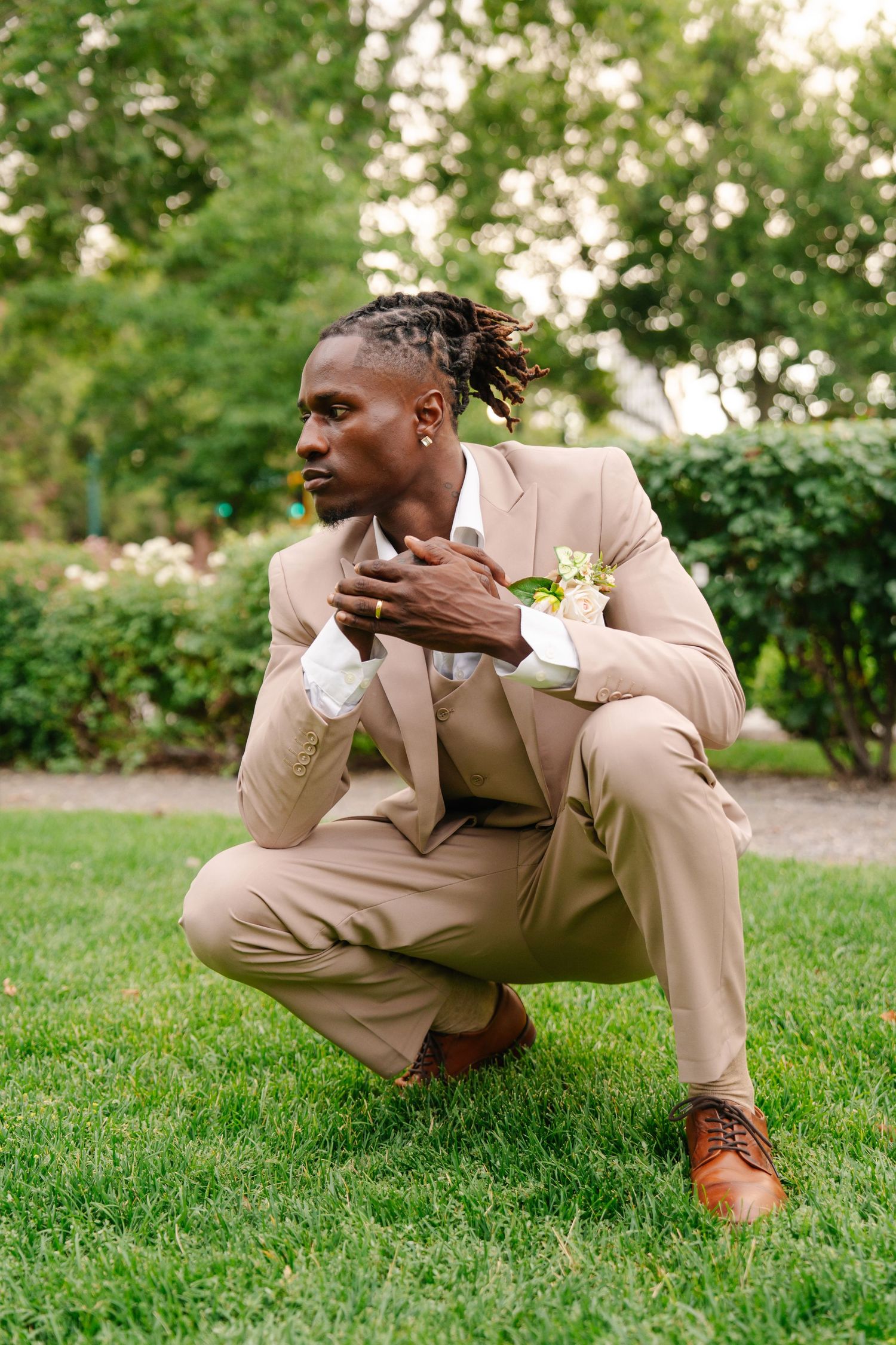 A well-dressed Black man in a beige suit with a floral boutonniere crouches on green grass in an outdoor garden setting.