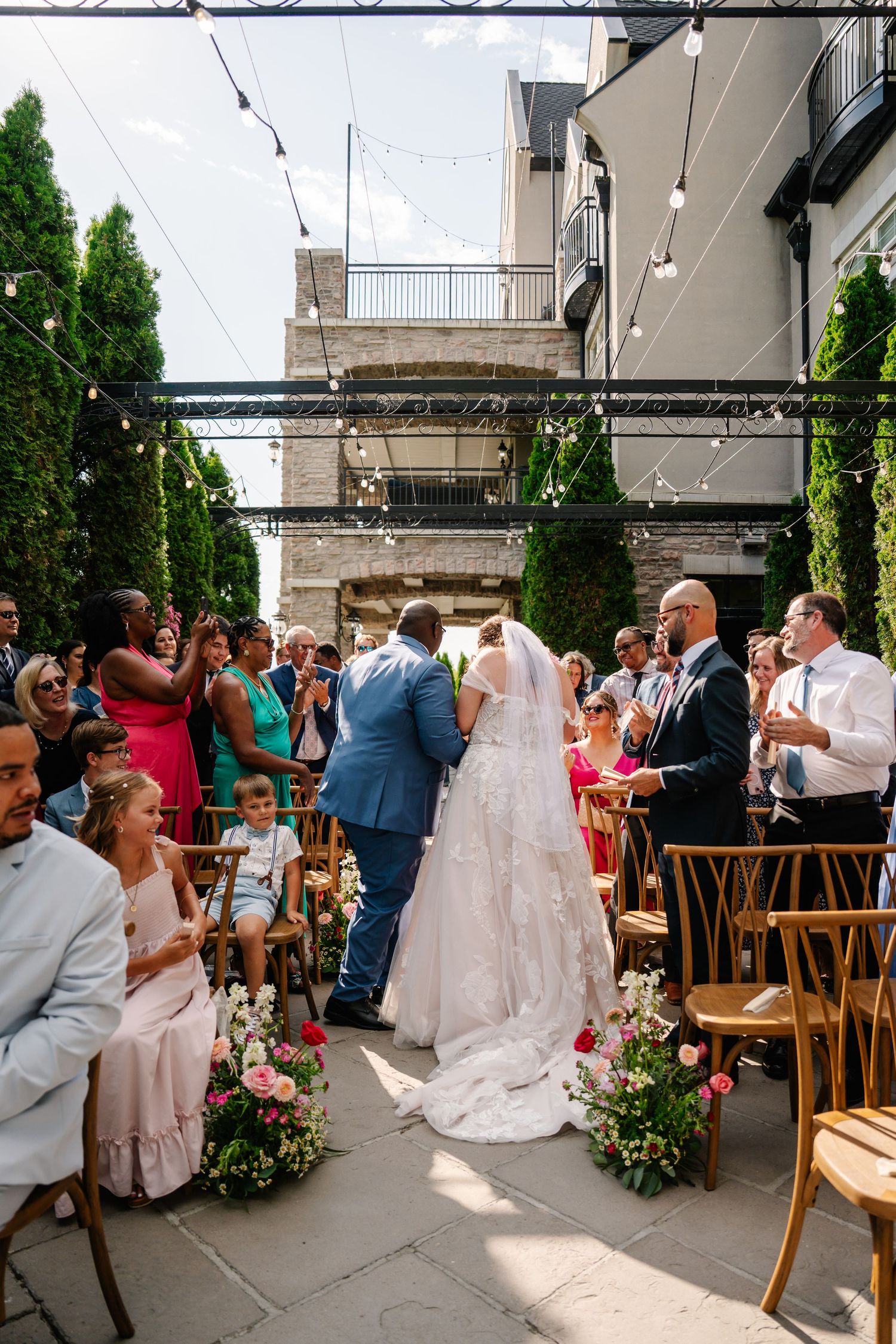 Bride and groom share a first kiss at an outdoor wedding ceremony with string lights and floral arrangements lining the aisle.