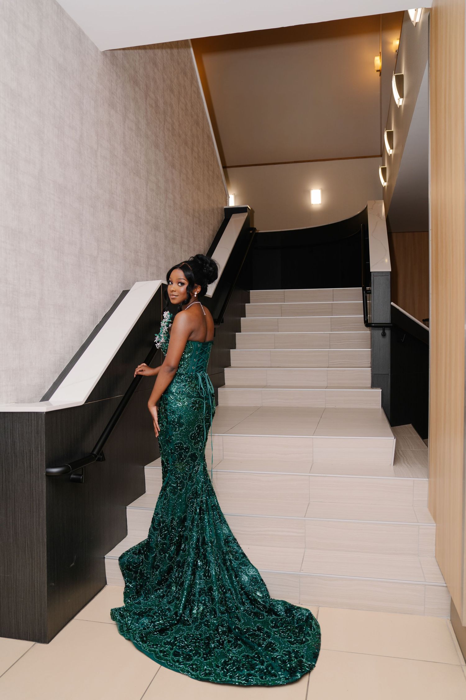 Woman in emerald green sequin mermaid gown with train poses elegantly on white marble staircase.