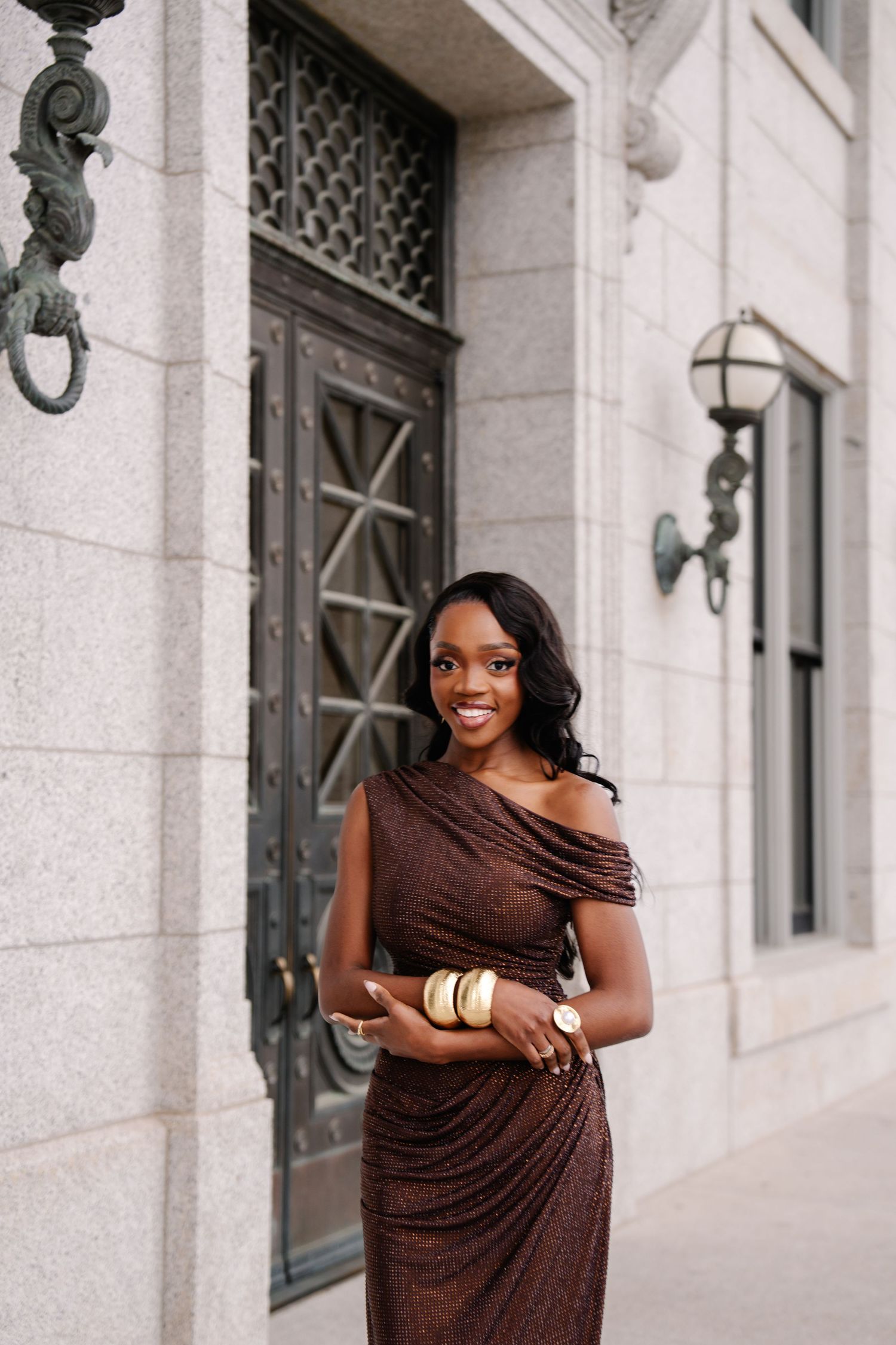 Smiling woman in brown sparkly off-shoulder gown with gold bracelets posing outside elegant stone building.