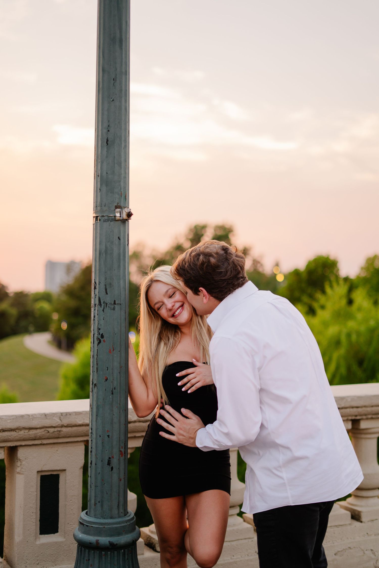 A couple shares a romantic moment at sunset near a lamppost, the man kissing the smiling woman in a black dress.