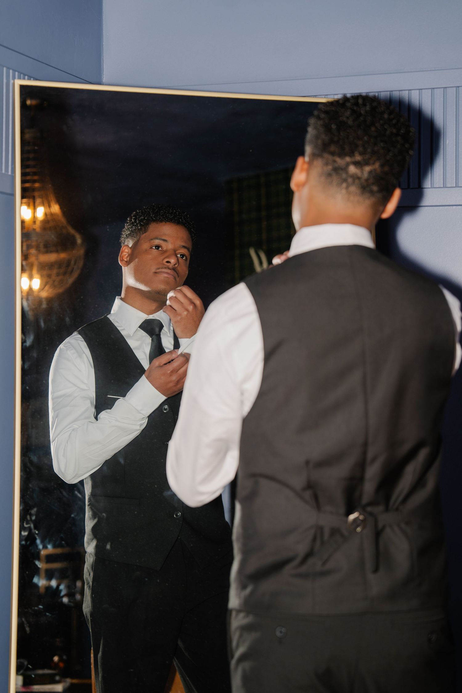 A well-dressed Black man in a black vest and white shirt adjusts his tie while looking in a mirror.