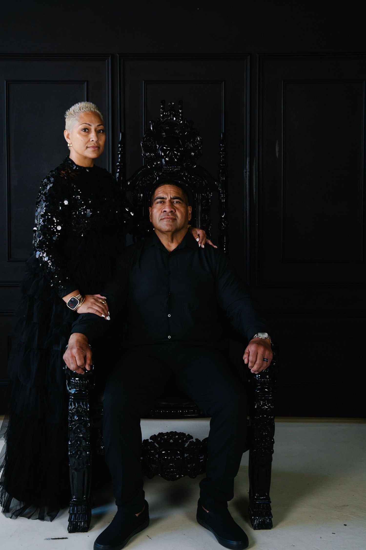 A couple dressed in elegant all-black attire poses regally, man seated on ornate black throne, woman standing beside him.