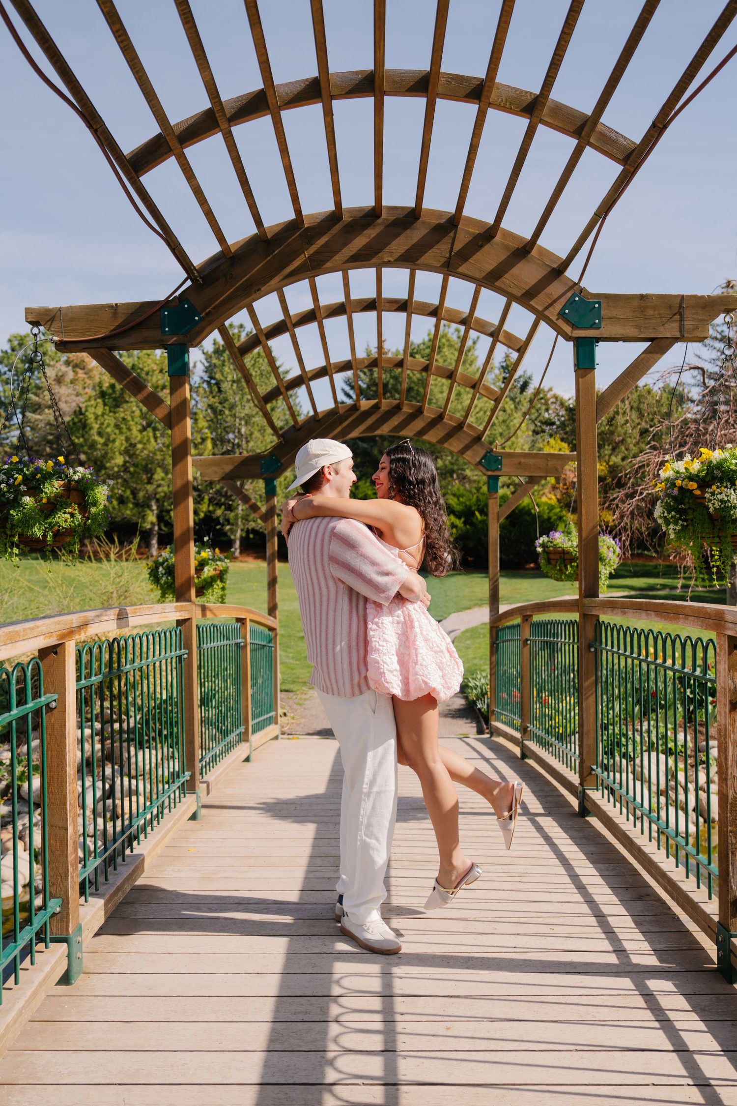 Couple embracing romantically on a wooden pergola bridge in a lush green park on a sunny day.