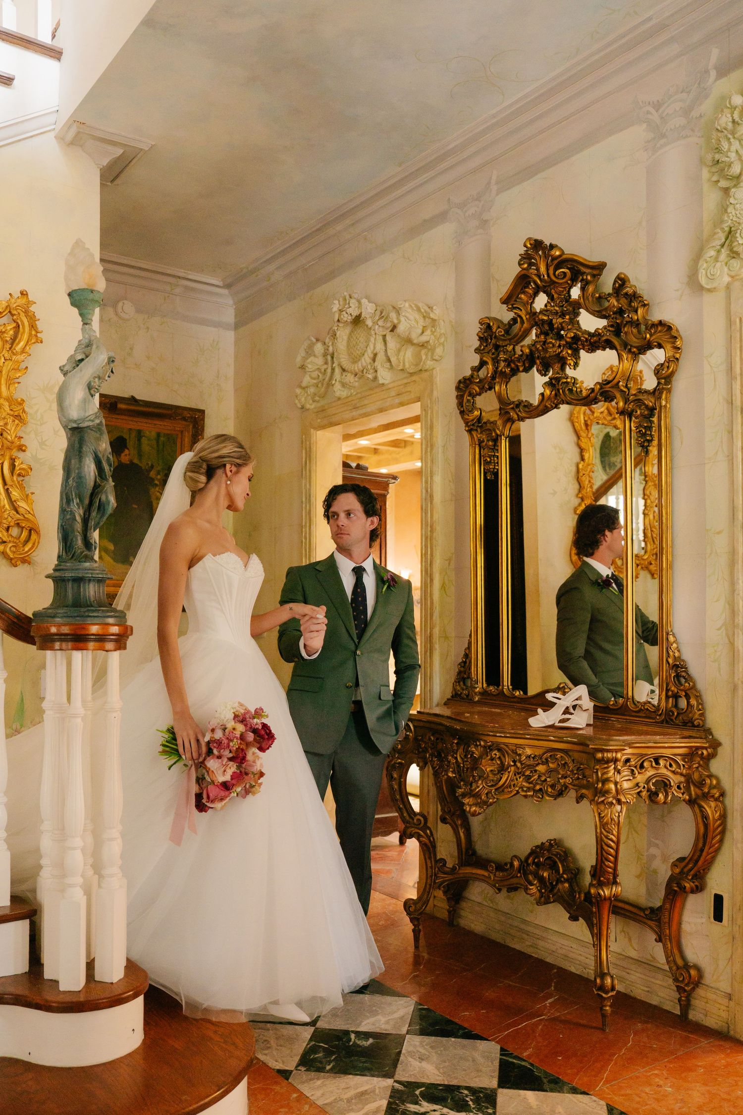 Wedding couple posing in an ornate hallway with gilded mirror and classical decor.