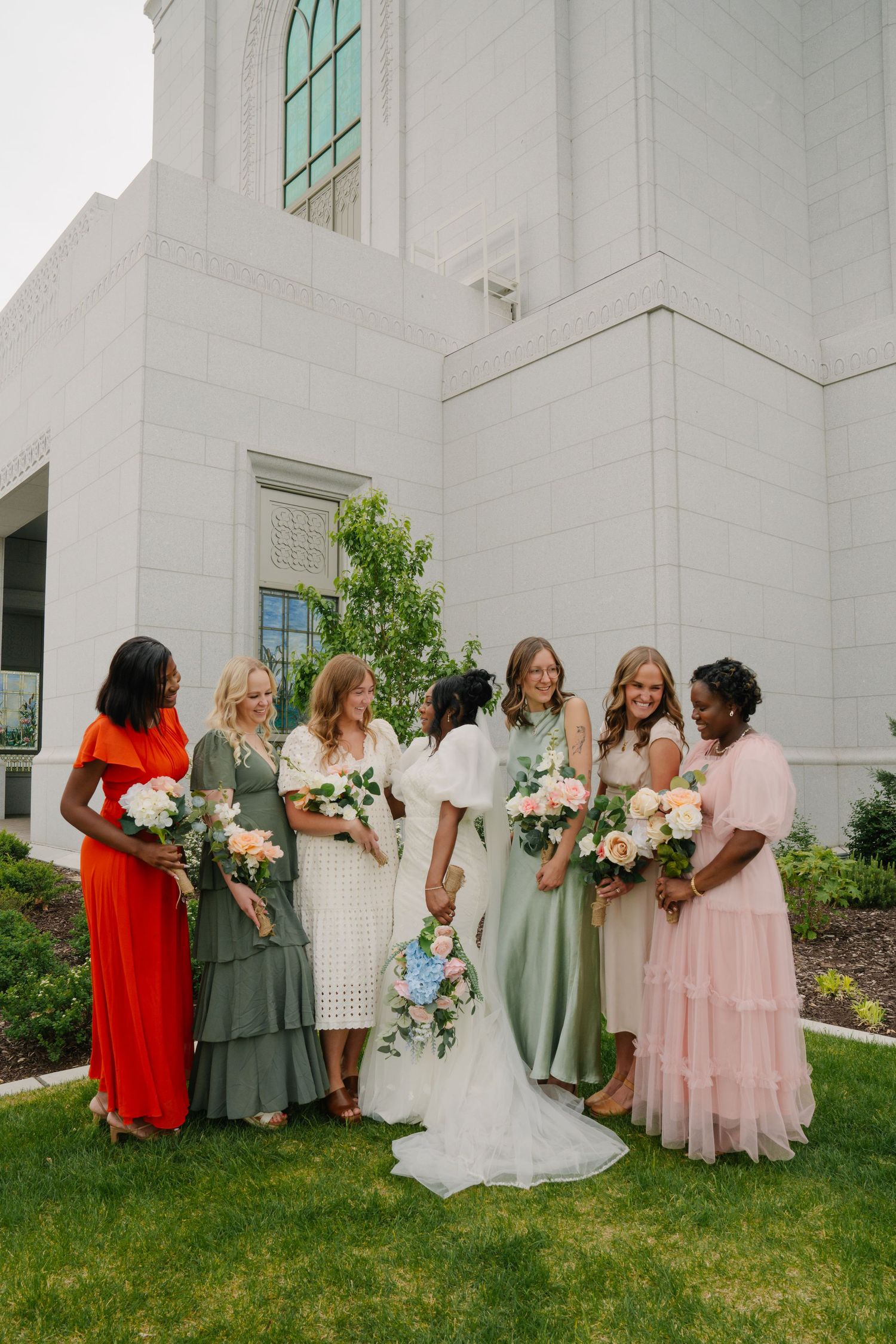 Bride and bridesmaids in colorful mismatched dresses holding bouquets outside a white stone wedding venue.