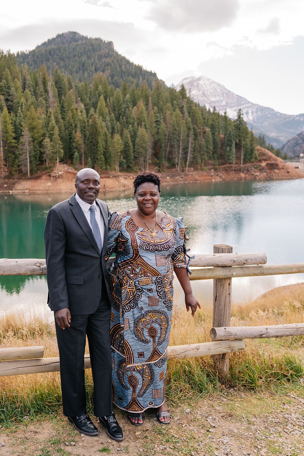 A couple poses together by a turquoise mountain lake with pine forests in the background.