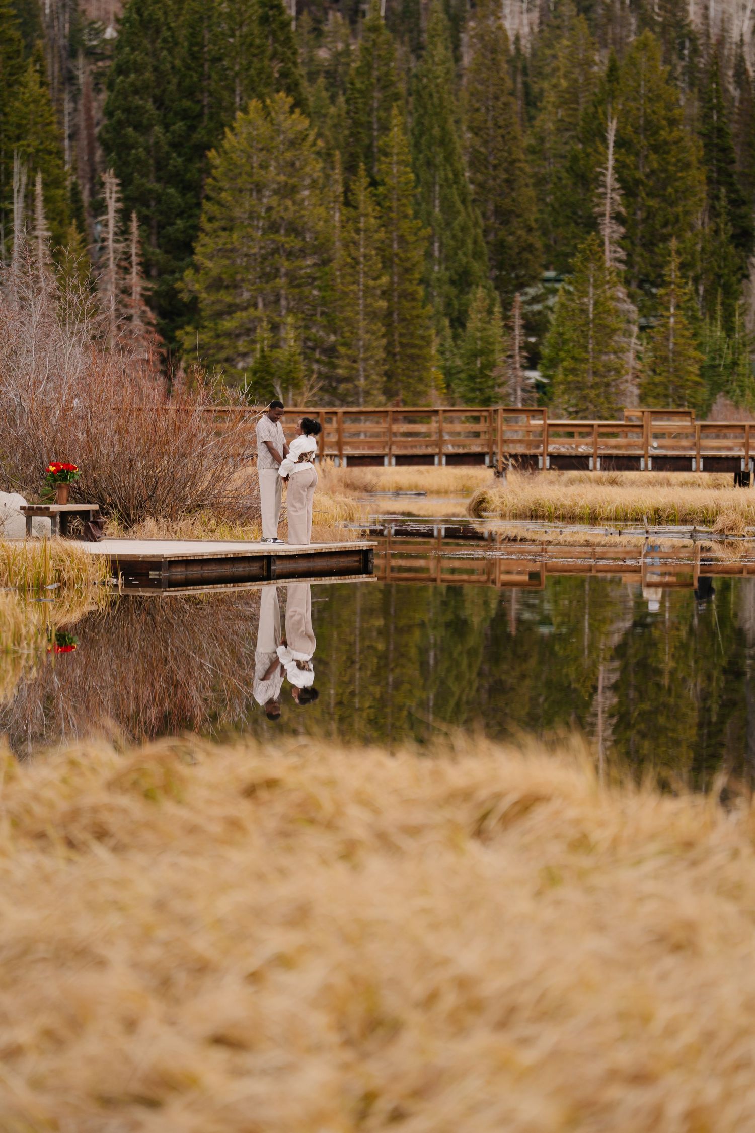 Couple embracing on a dock reflected in a calm mountain lake surrounded by pine trees and golden grass.