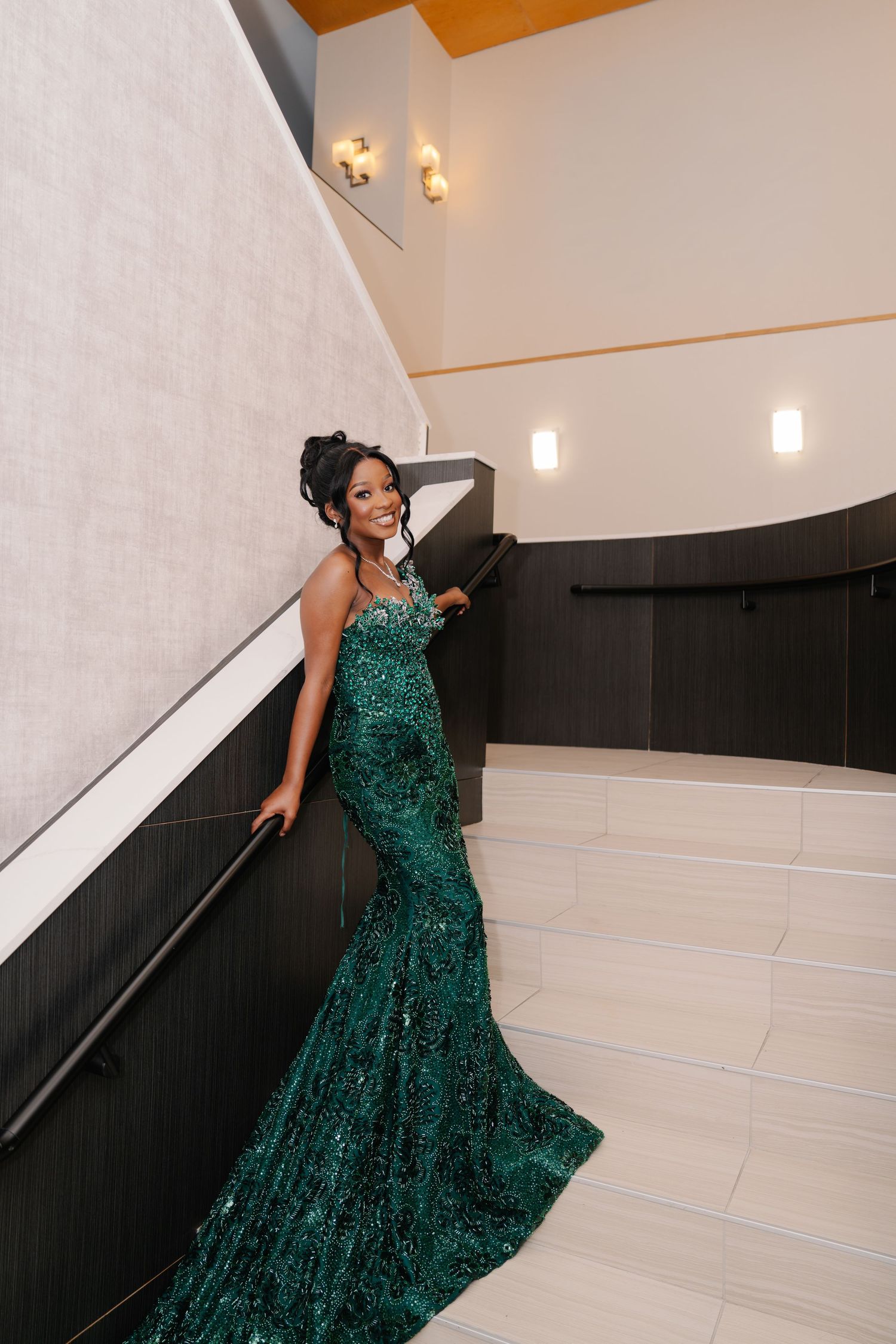 A woman in a sparkling emerald green sequined mermaid gown poses elegantly on a white staircase.