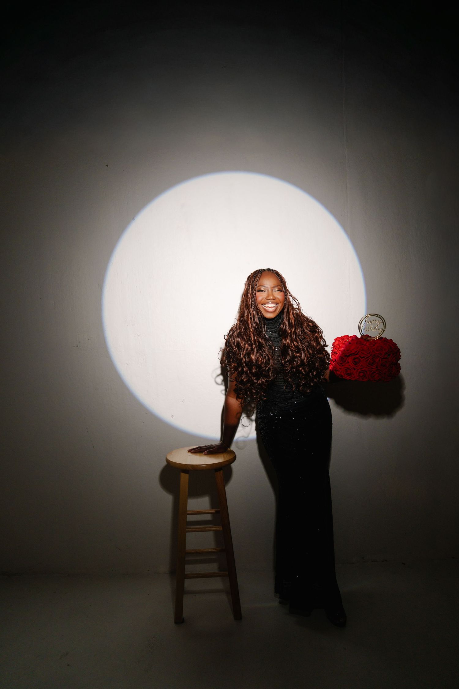 Smiling woman in black sequin dress holding red roses, posing beside wooden stool under spotlight on gray background.