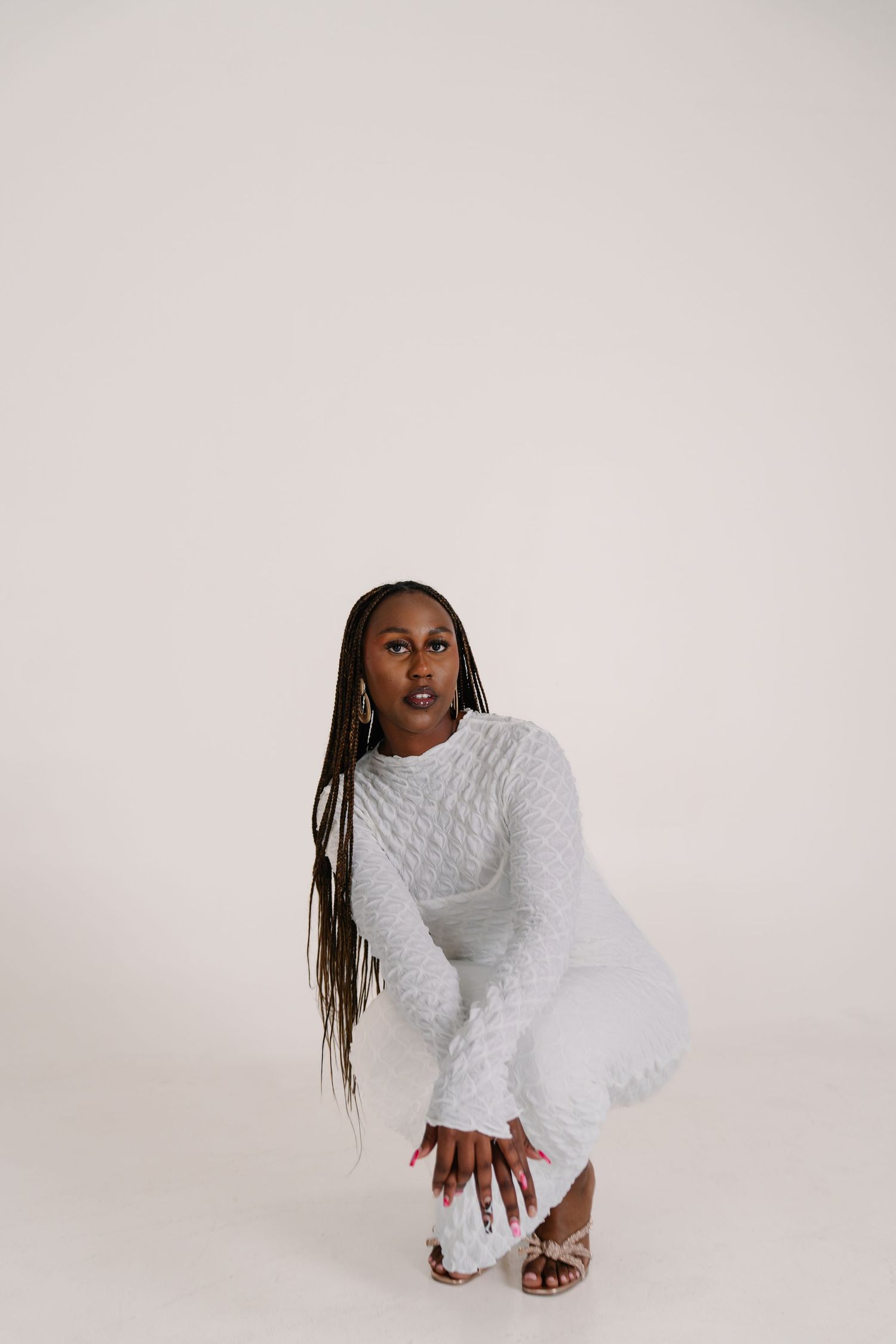 A young Black woman with long braids crouches in a white knit outfit against a light background.