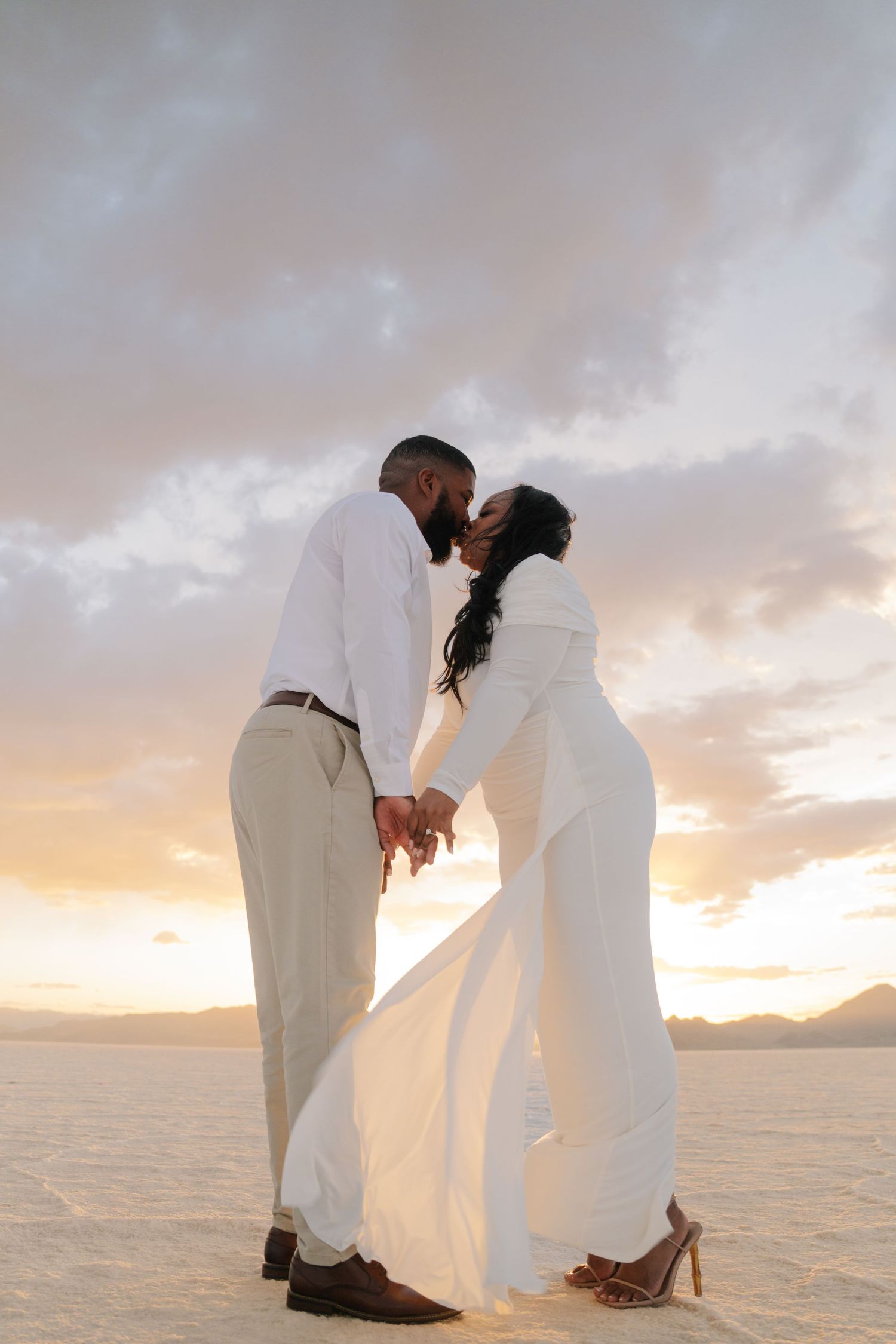 Couple in white attire holding hands and kissing at sunset on white salt flats.