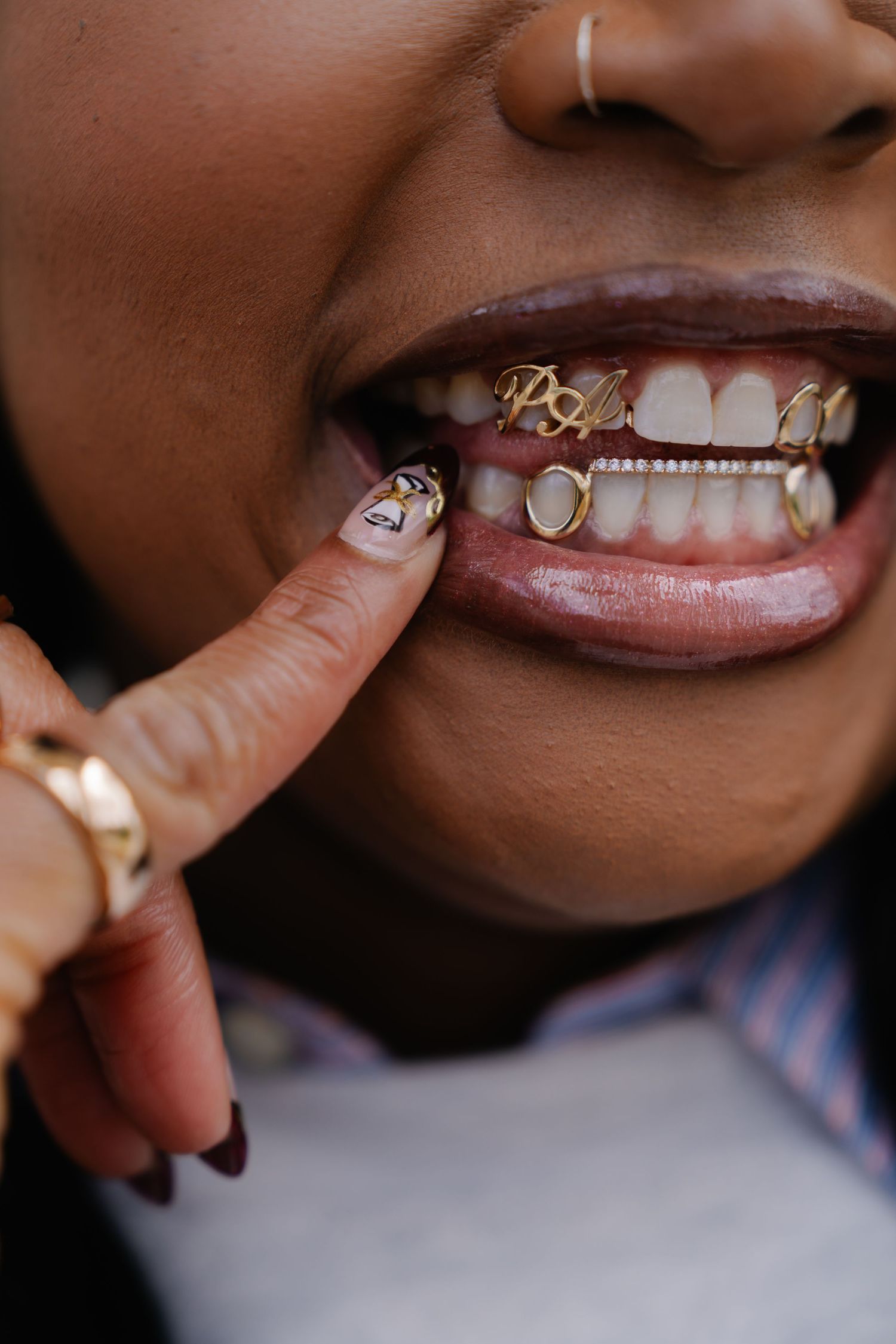 Close-up of a woman showing gold grillz on her teeth, wearing rings and decorative nail art.