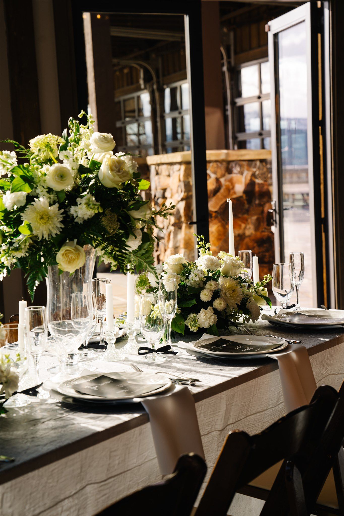 Elegant wedding reception table setting with white floral centerpieces and crystal glassware in natural light.
