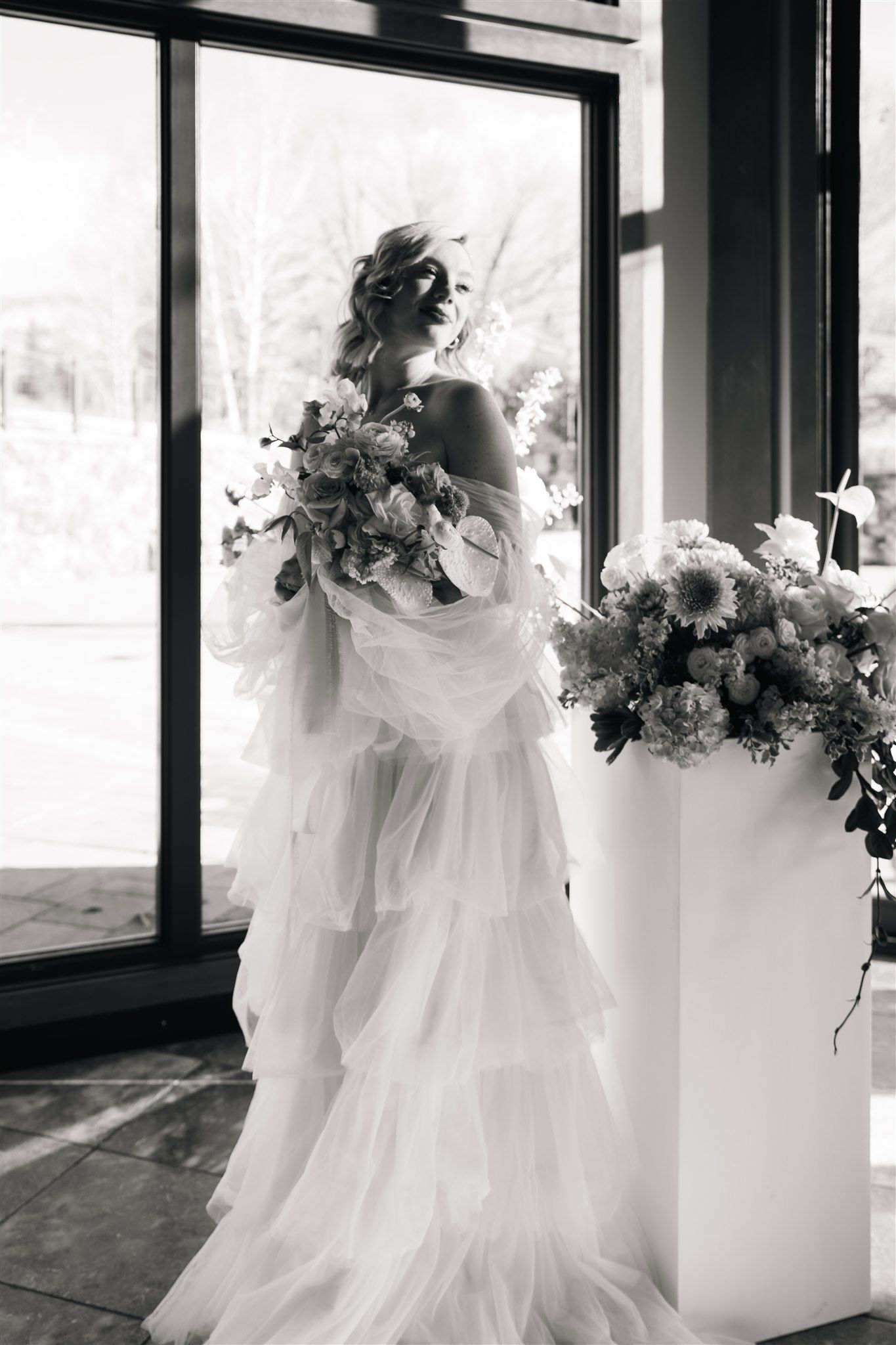 Elegant black and white photo of bride with cascading bouquet and ruffled gown near floor-to-ceiling windows