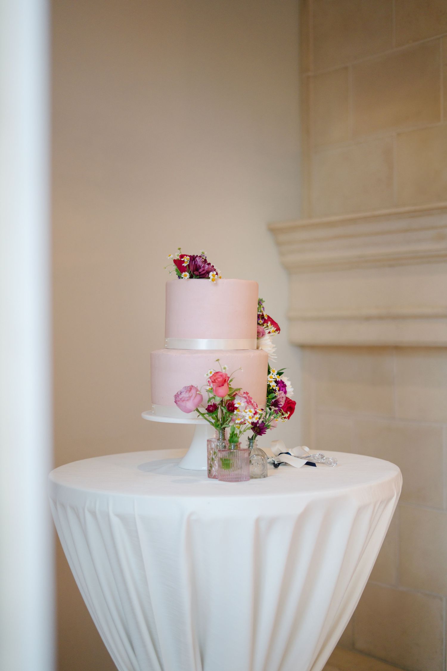 Elegant two-tier pink wedding cake decorated with fresh flowers on a white tablecloth.