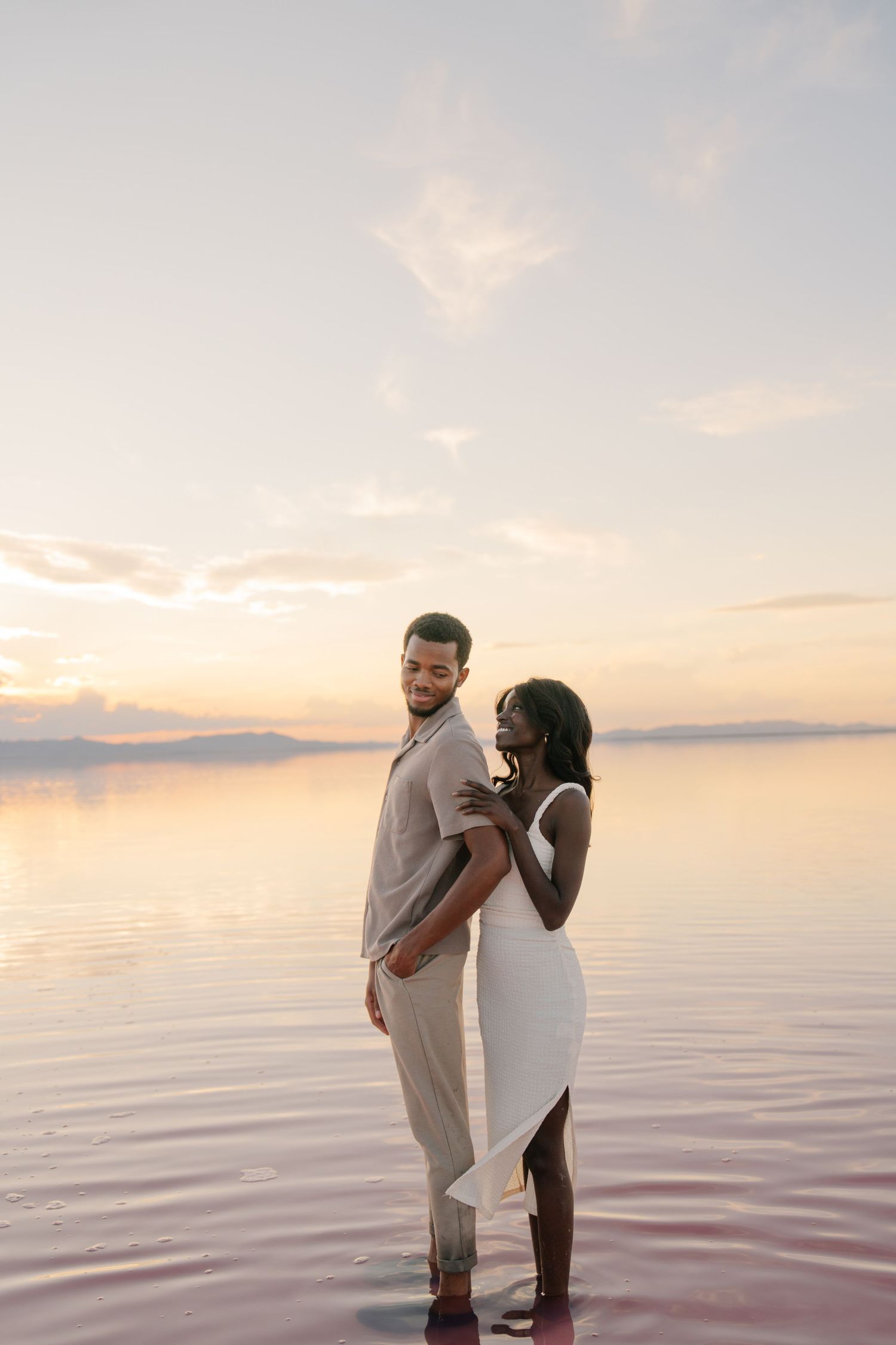 A romantic couple embraces while standing in pink-hued waters during golden hour.