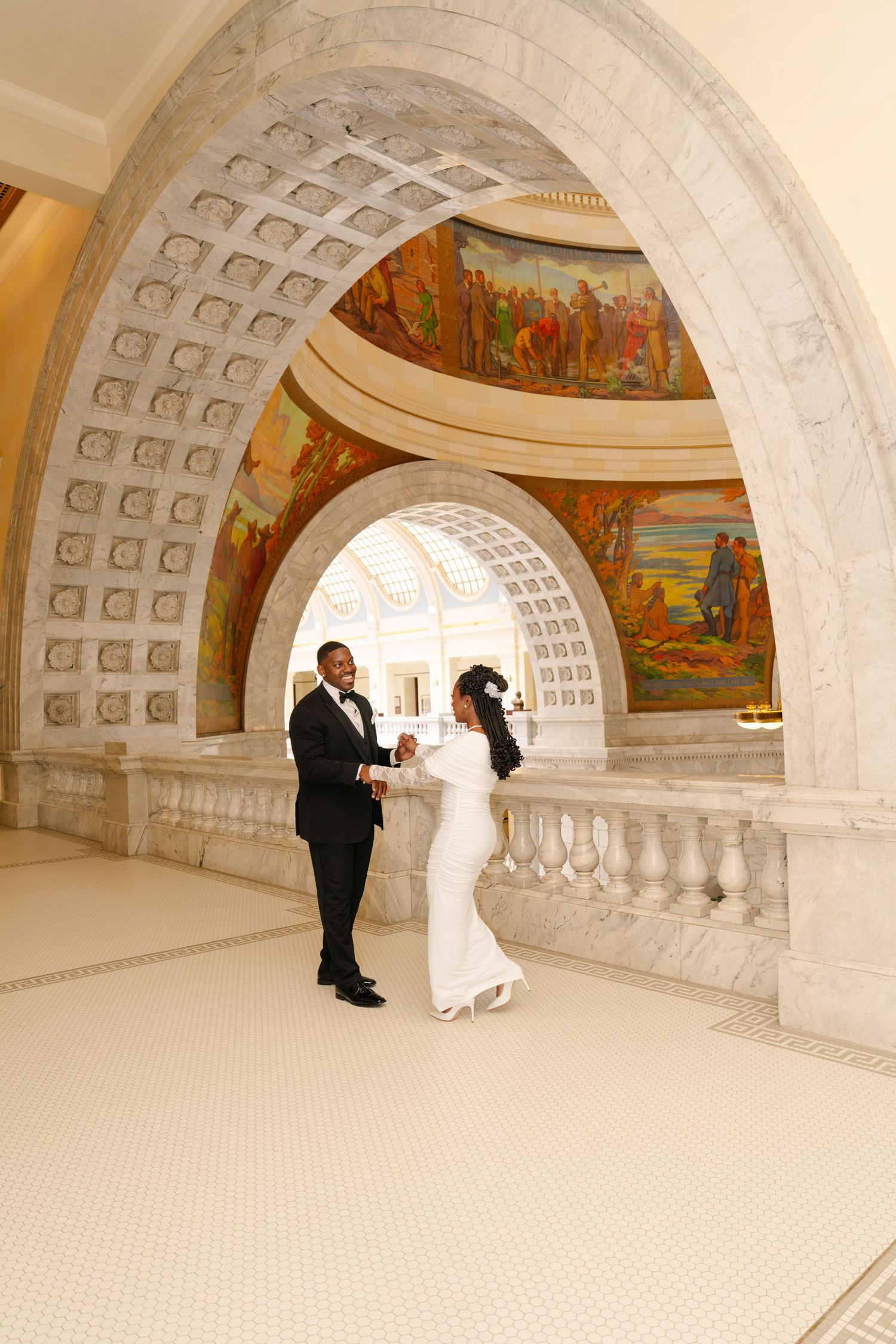 Newlywed couple dancing under ornate marble arches with colorful murals in an elegant government building.